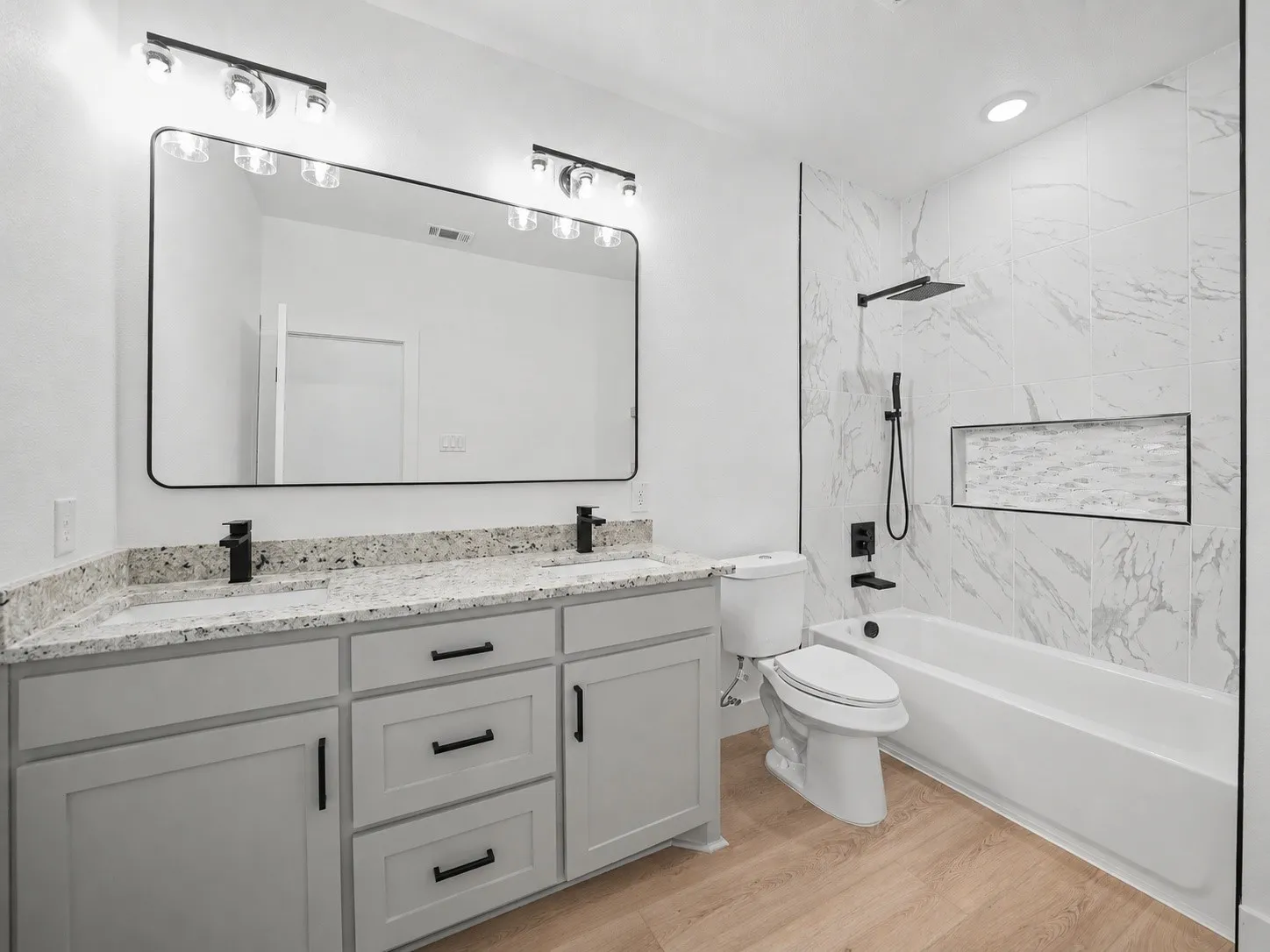 Bathroom featuring a double vanity with granite-style countertops, light wood-finish flooring, a full bathtub with a shower niche, and matte black fixtures