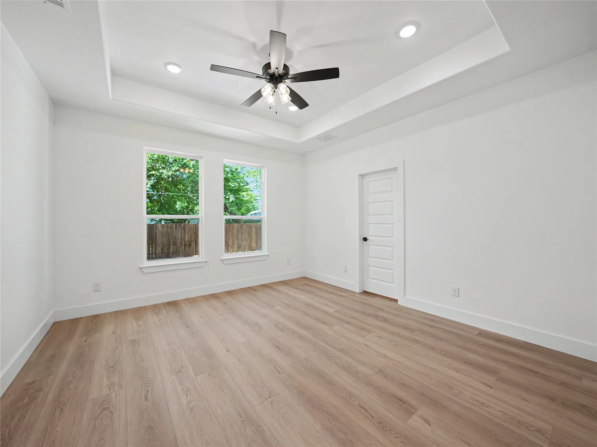 Spacious room featuring wood-finish flooring, a tray ceiling with recessed lighting and a ceiling fan, and two large windows offering natural light