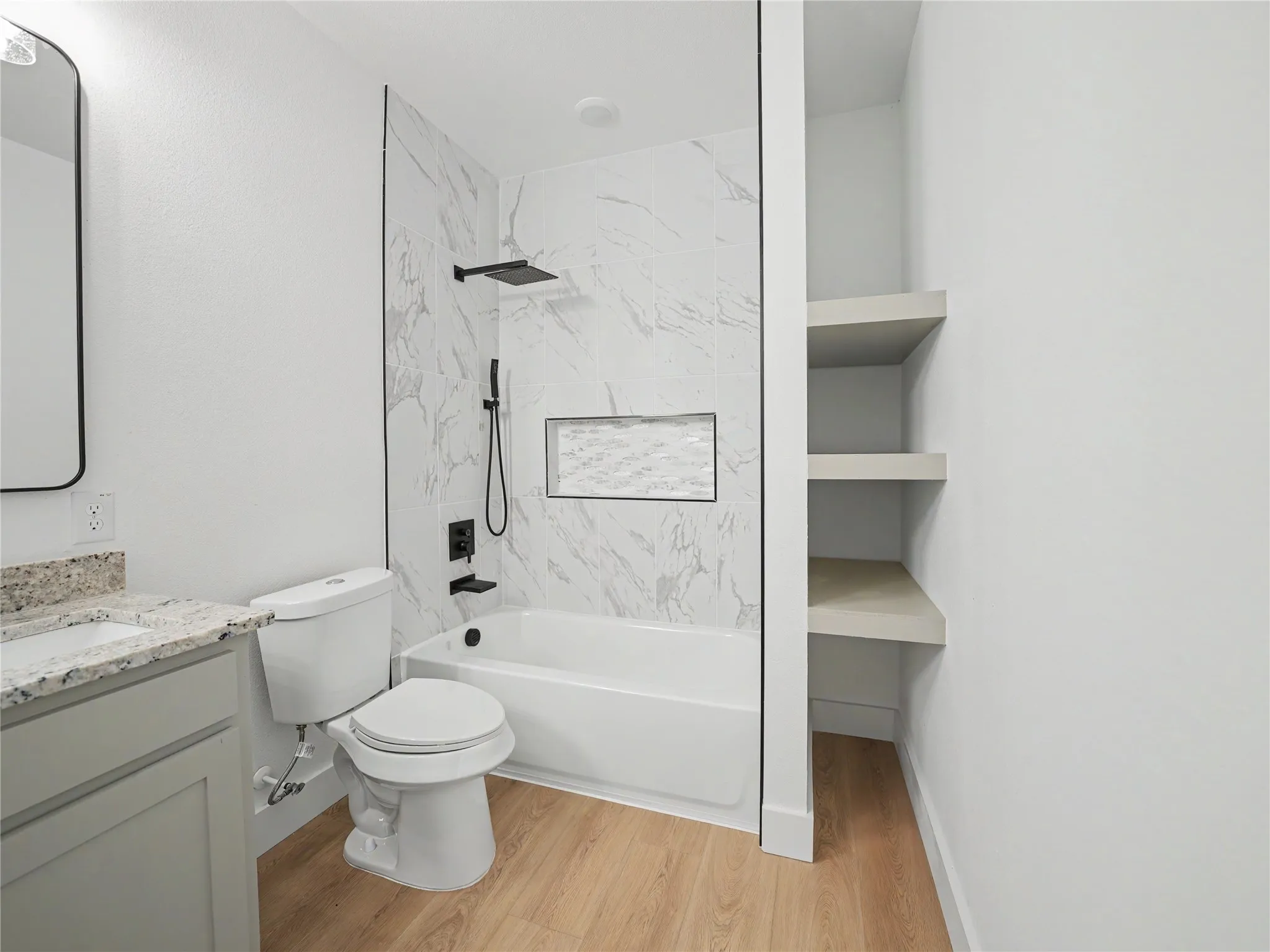 Contemporary bathroom featuring a white tub with marble-patterned tile surround, matte black shower fixtures, a built-in shower niche, and light wood-finish flooring