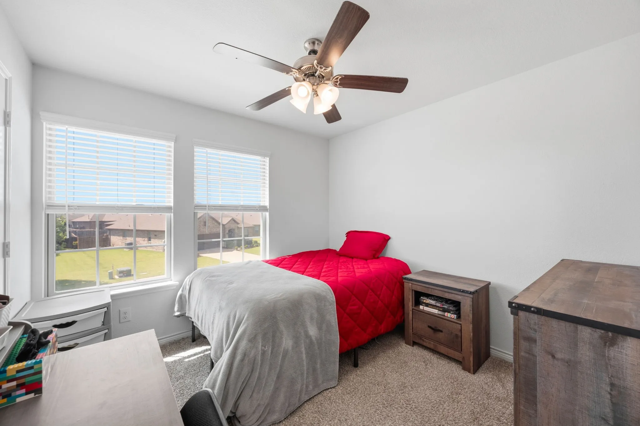 Bright bedroom featuring two windows with horizontal blinds, a ceiling fan with light kit, textured carpet flooring, and white walls