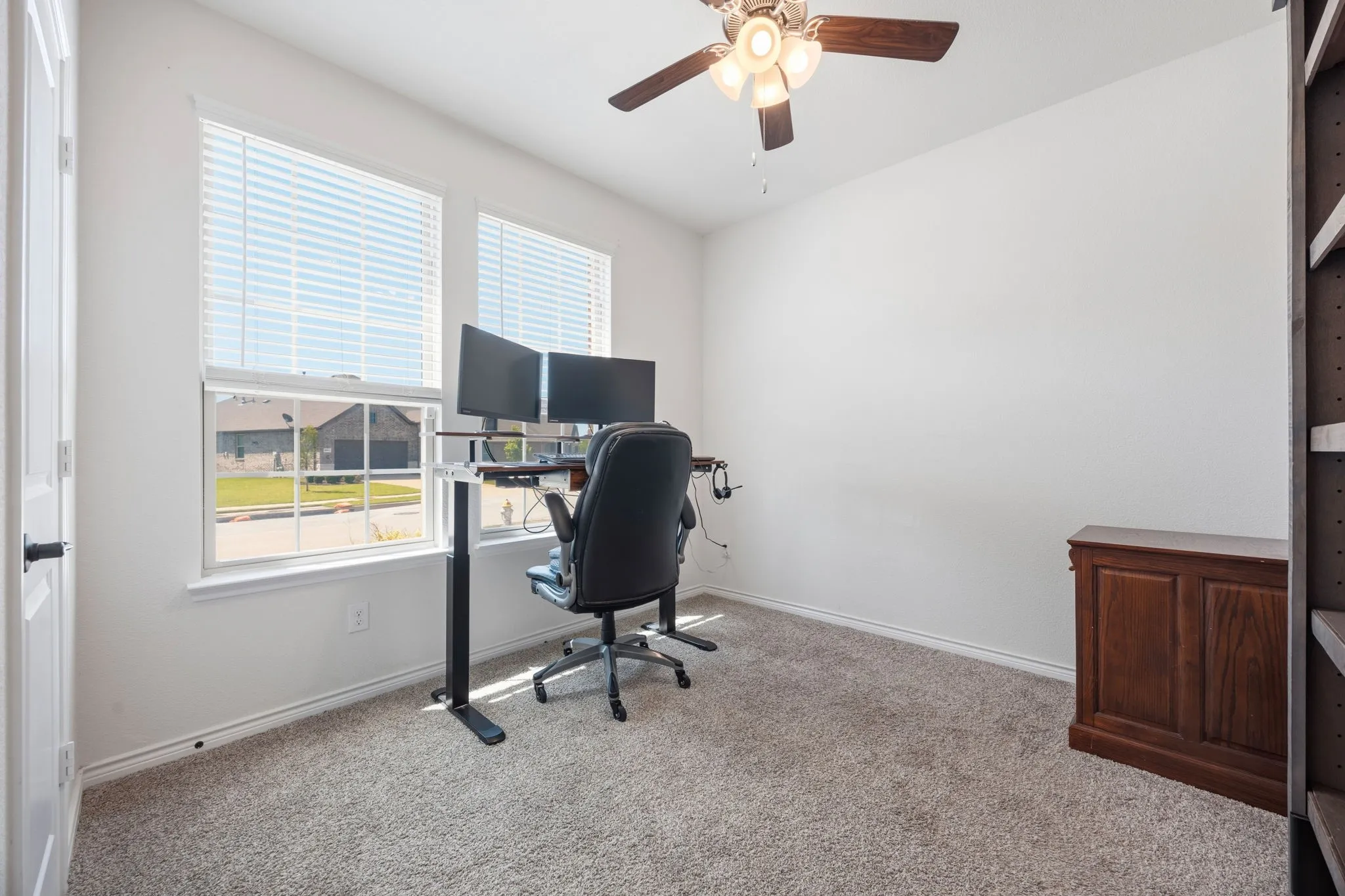 Versatile room featuring two large windows with blinds, plush carpet flooring, a ceiling fan with integrated lighting, and white painted walls