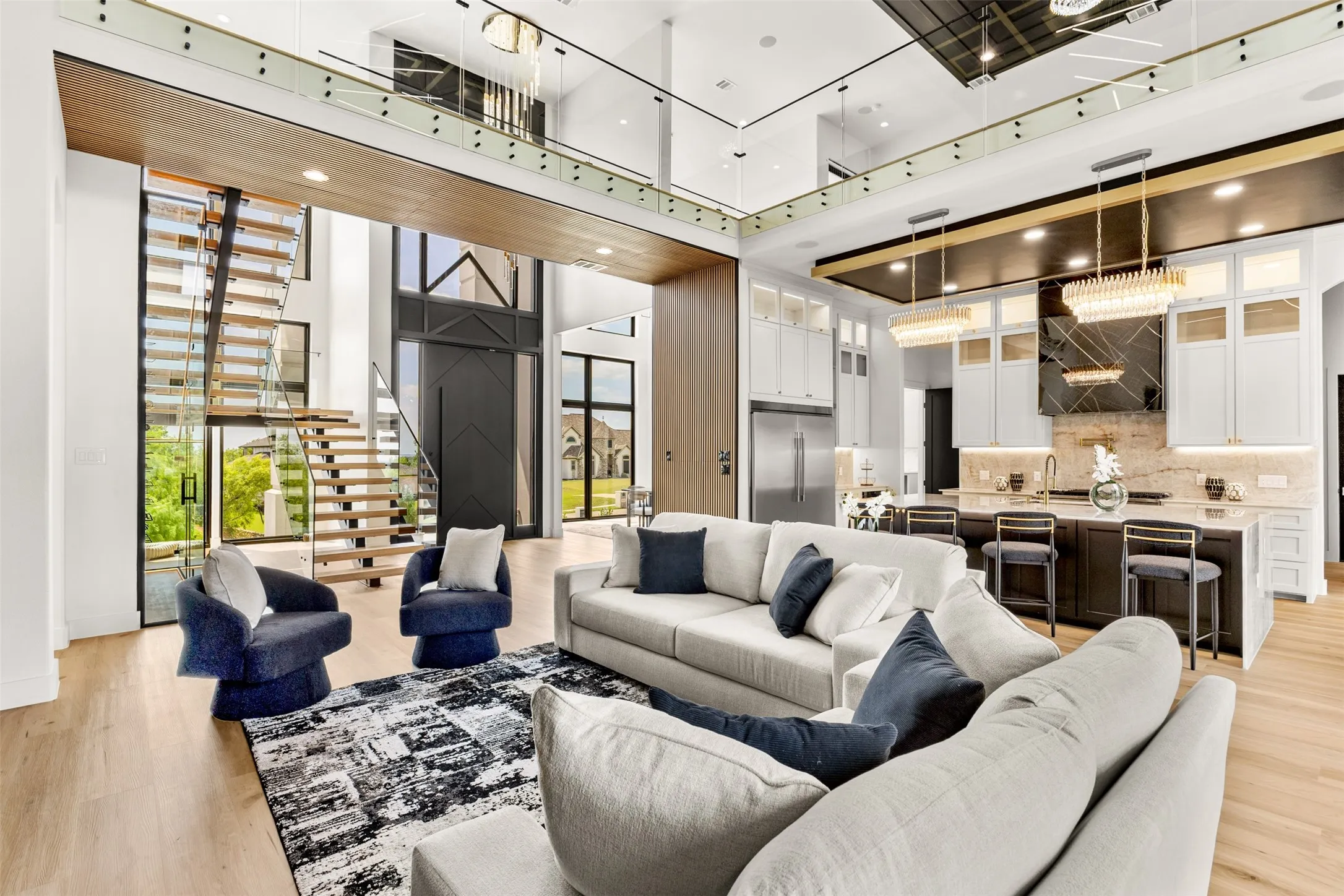 Open-concept interior featuring light wood-finish flooring, a contemporary staircase with glass railings, a chef's kitchen with a large island and pendant lighting, a two-story foyer with a dark paneled door, and a second-story interior balcony