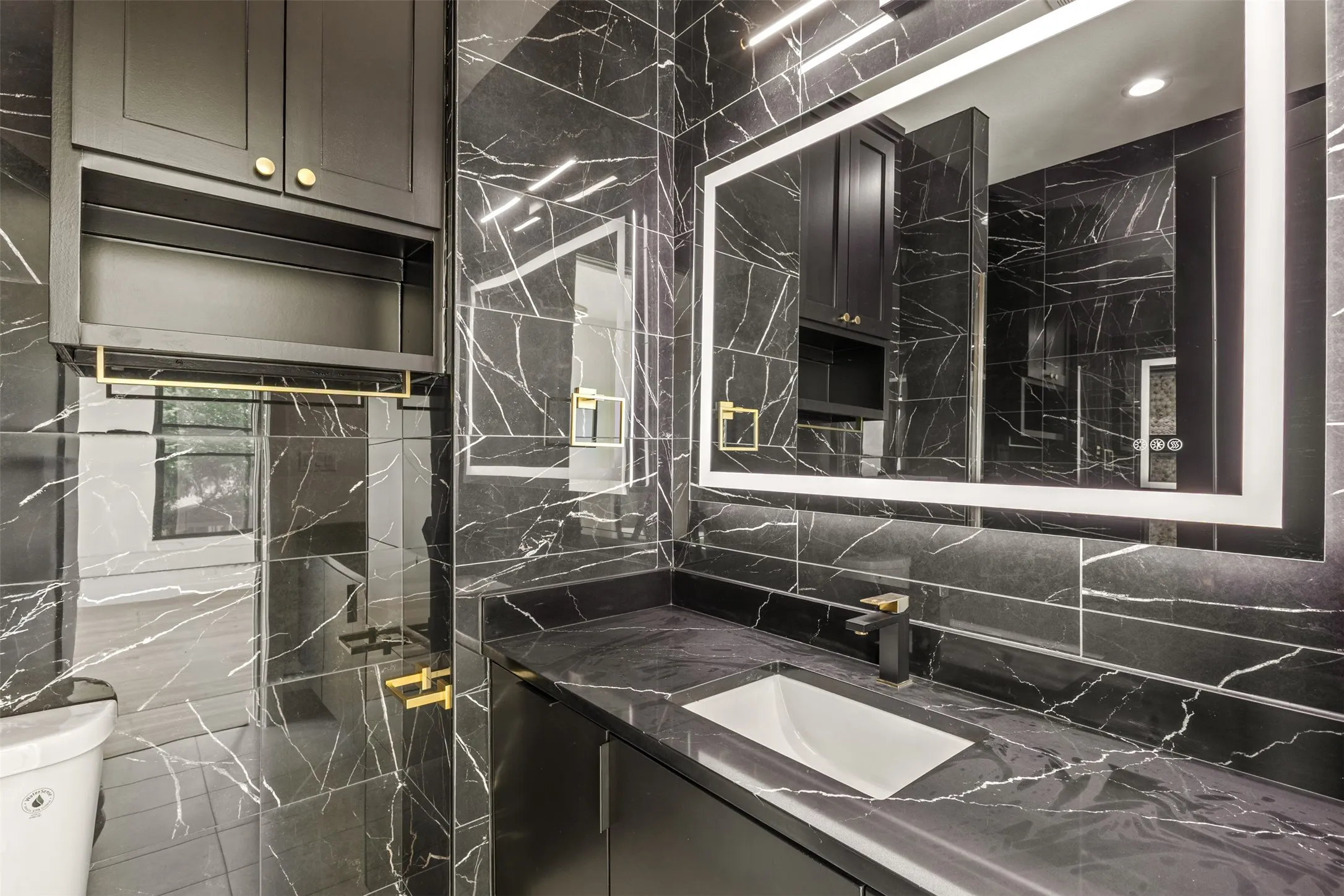 Bathroom featuring black vein-patterned wall and floor tile, a dark-toned vanity with an integrated sink, a large backlit mirror, and a black and gold-toned faucet