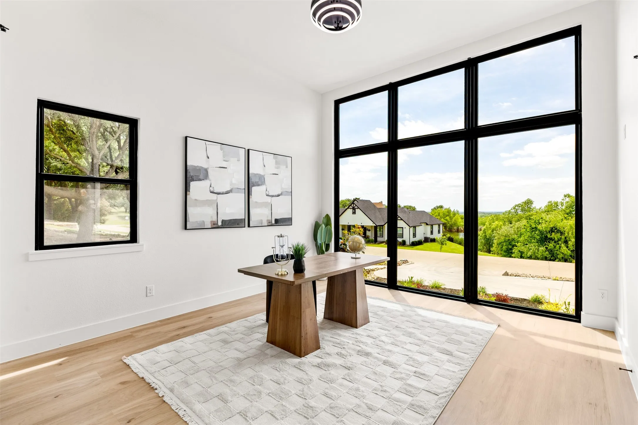 Expansive floor-to-ceiling windows with black frames, light wood-finish flooring, white walls, and a modern ceiling light fixture