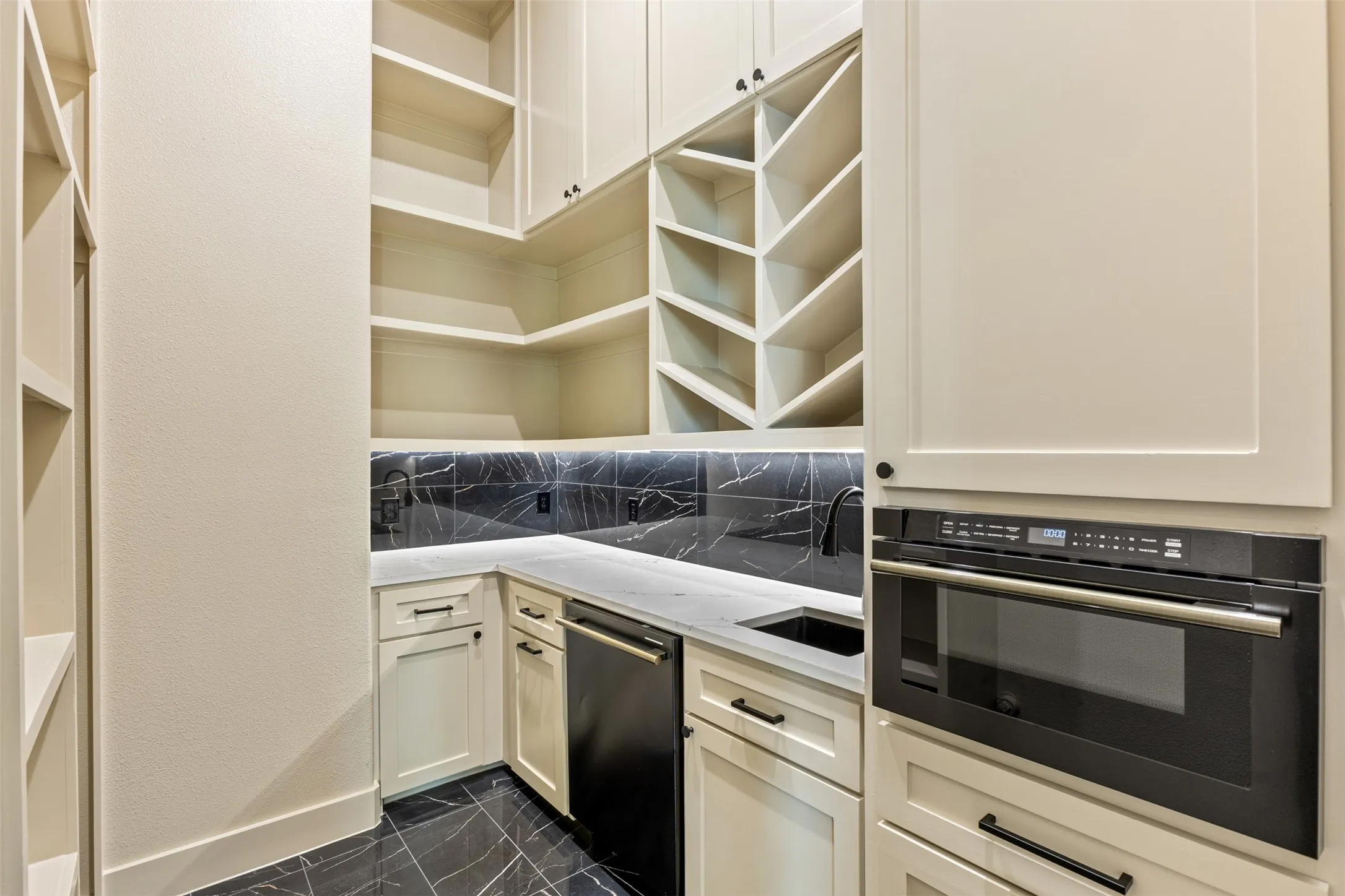 Dedicated pantry space featuring built-in shelving, integrated microwave, black marble-look tile backsplash, under-cabinet lighting, and a black sink with a matte black faucet