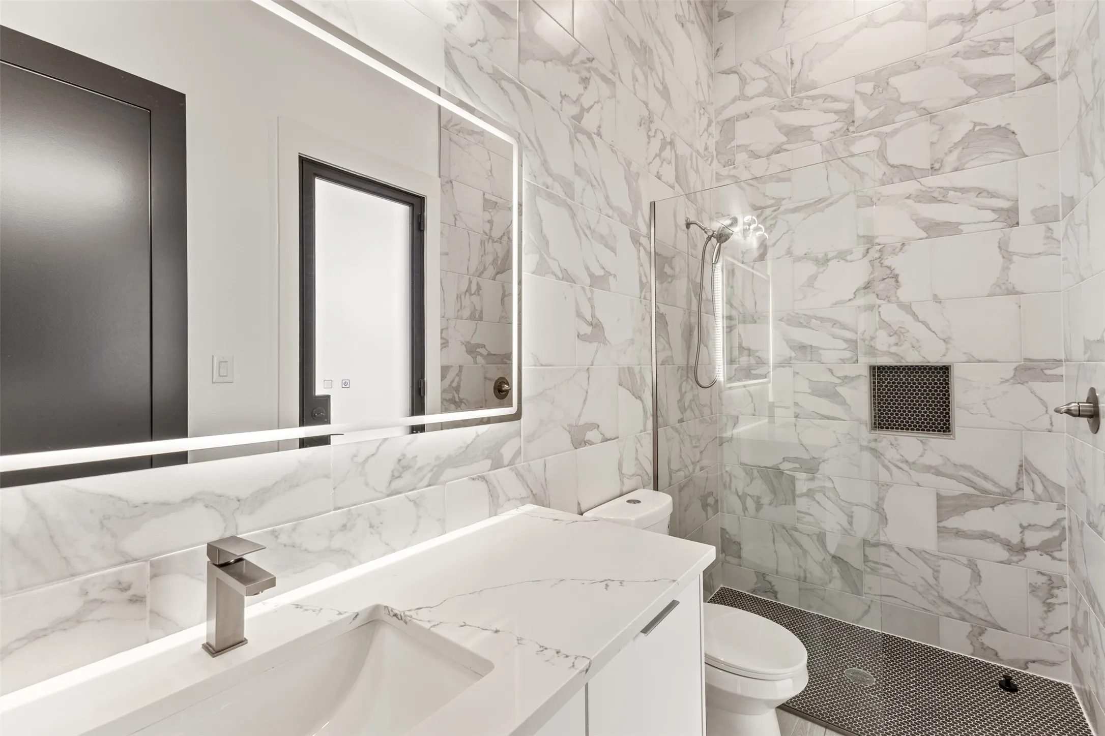 Modern bathroom featuring a white vanity with an integrated sink, a large illuminated mirror, and a walk-in shower with stone-look tile walls, a built-in niche, and dark penny tile flooring