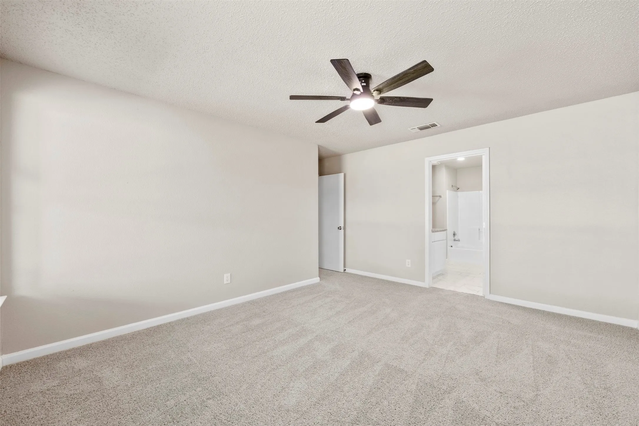 Spacious room featuring neutral wall paint, light-colored carpet flooring, a dark wood-finish ceiling fan with integrated lighting, and white baseboards