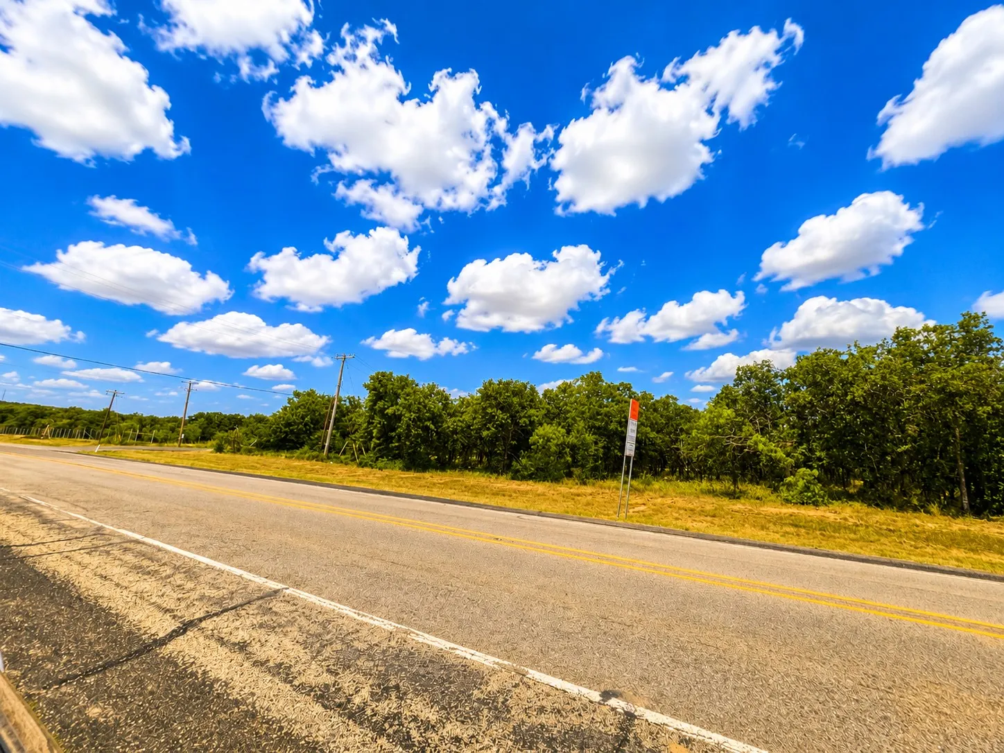 Paved road with a clear double yellow line, bordering a natural landscape featuring mature trees and open land