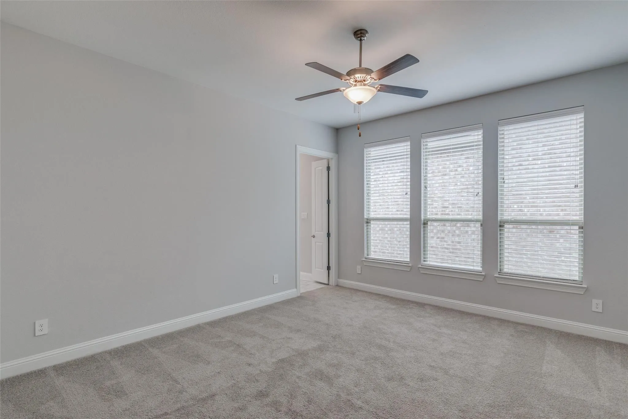 Neutral-toned room featuring a ceiling fan with integrated lighting, three large windows with horizontal blinds, gray carpet flooring, and white baseboards