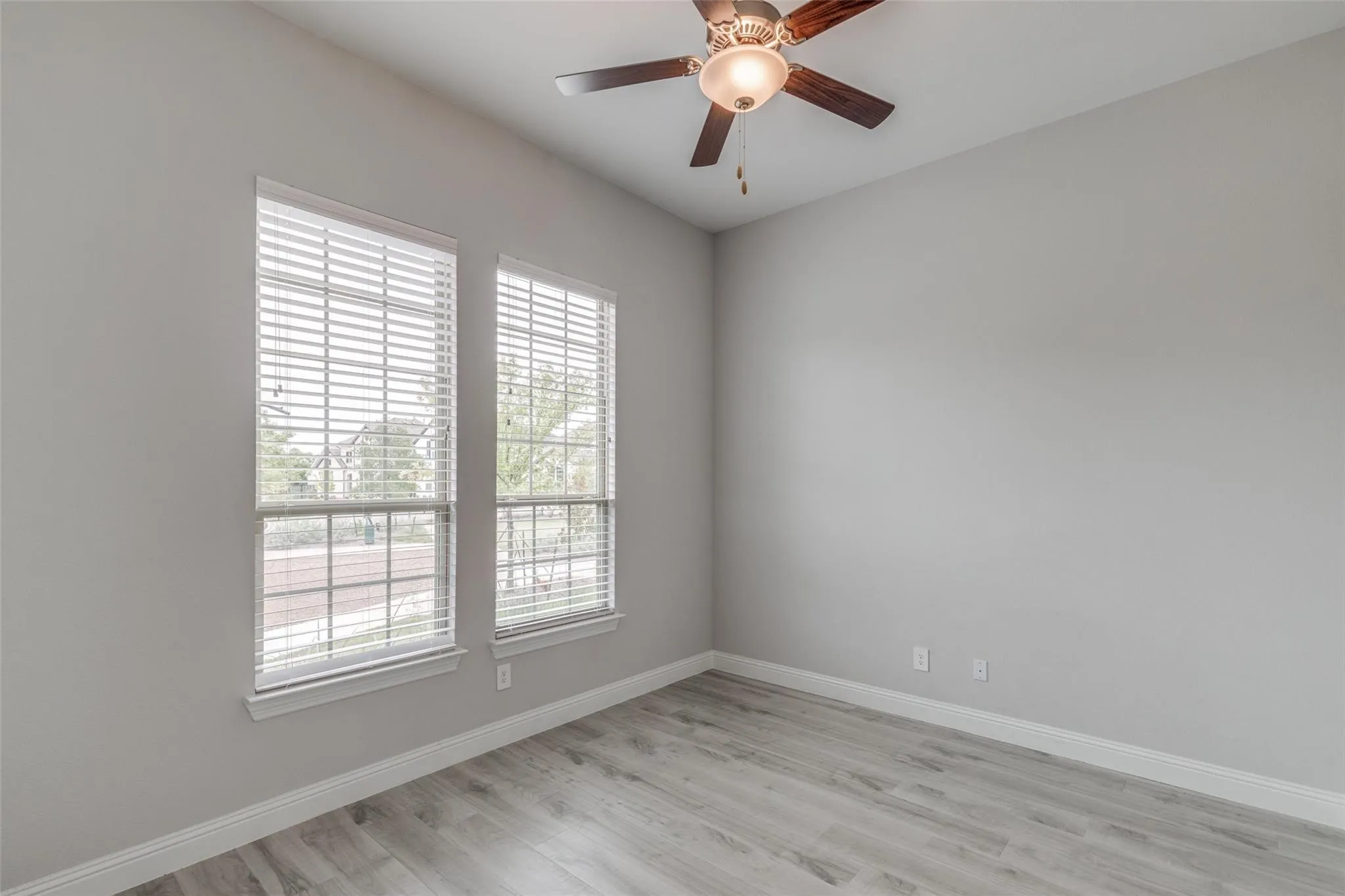 Room featuring two windows with blinds, wood-finish flooring, light grey walls, white baseboards, and a ceiling fan with light fixture