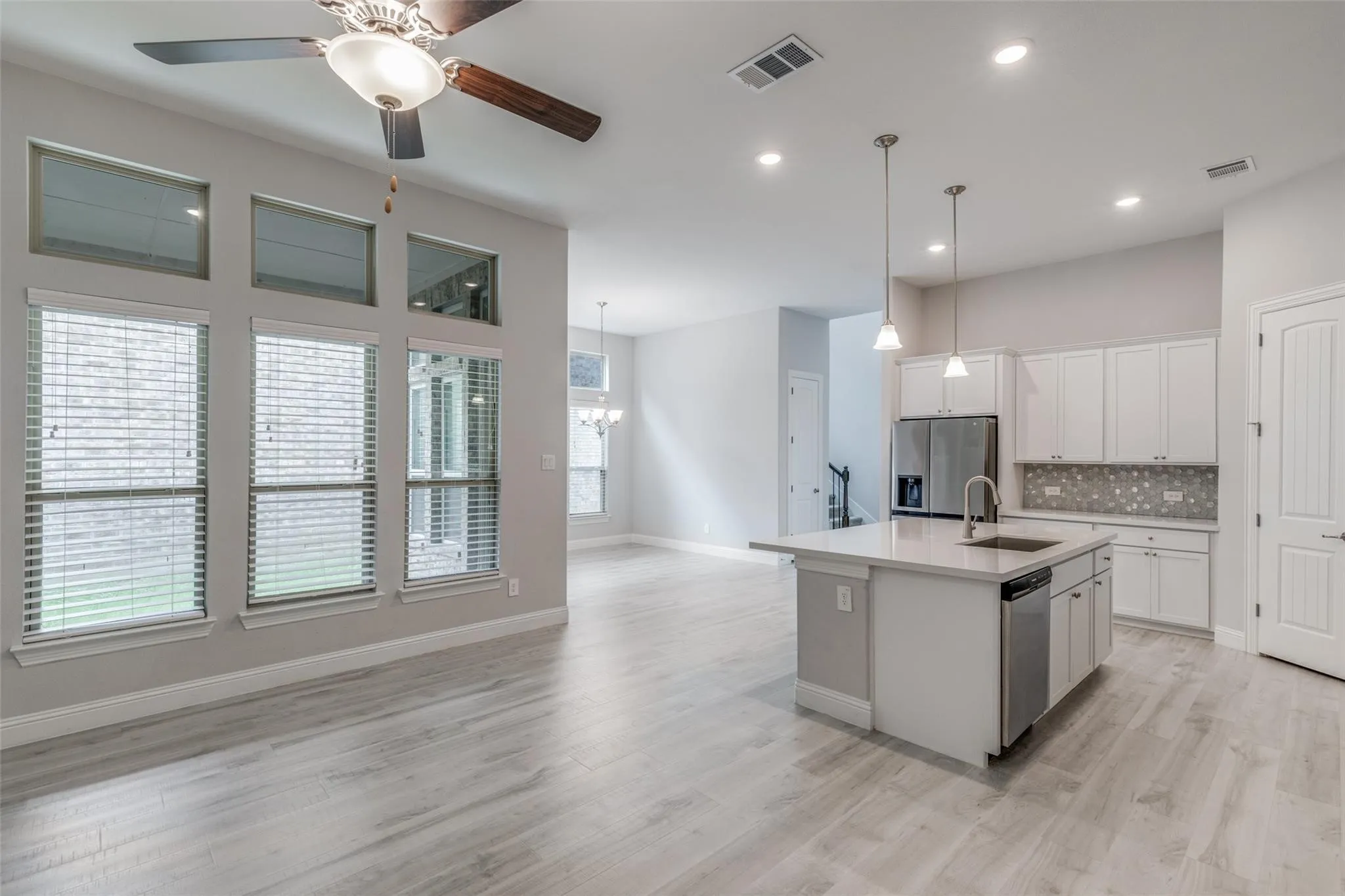 Open-concept living space featuring wood-finish flooring, a kitchen island with pendant lighting, stainless steel appliances, white cabinetry, and a full wall of windows with upper transom windows