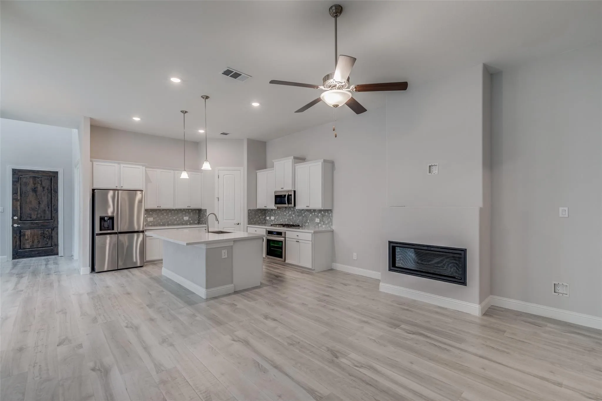 Open-concept living area featuring wood-finish flooring, a contemporary kitchen with white cabinetry and an island, stainless steel appliances, a tile backsplash, and a recessed electric fireplace