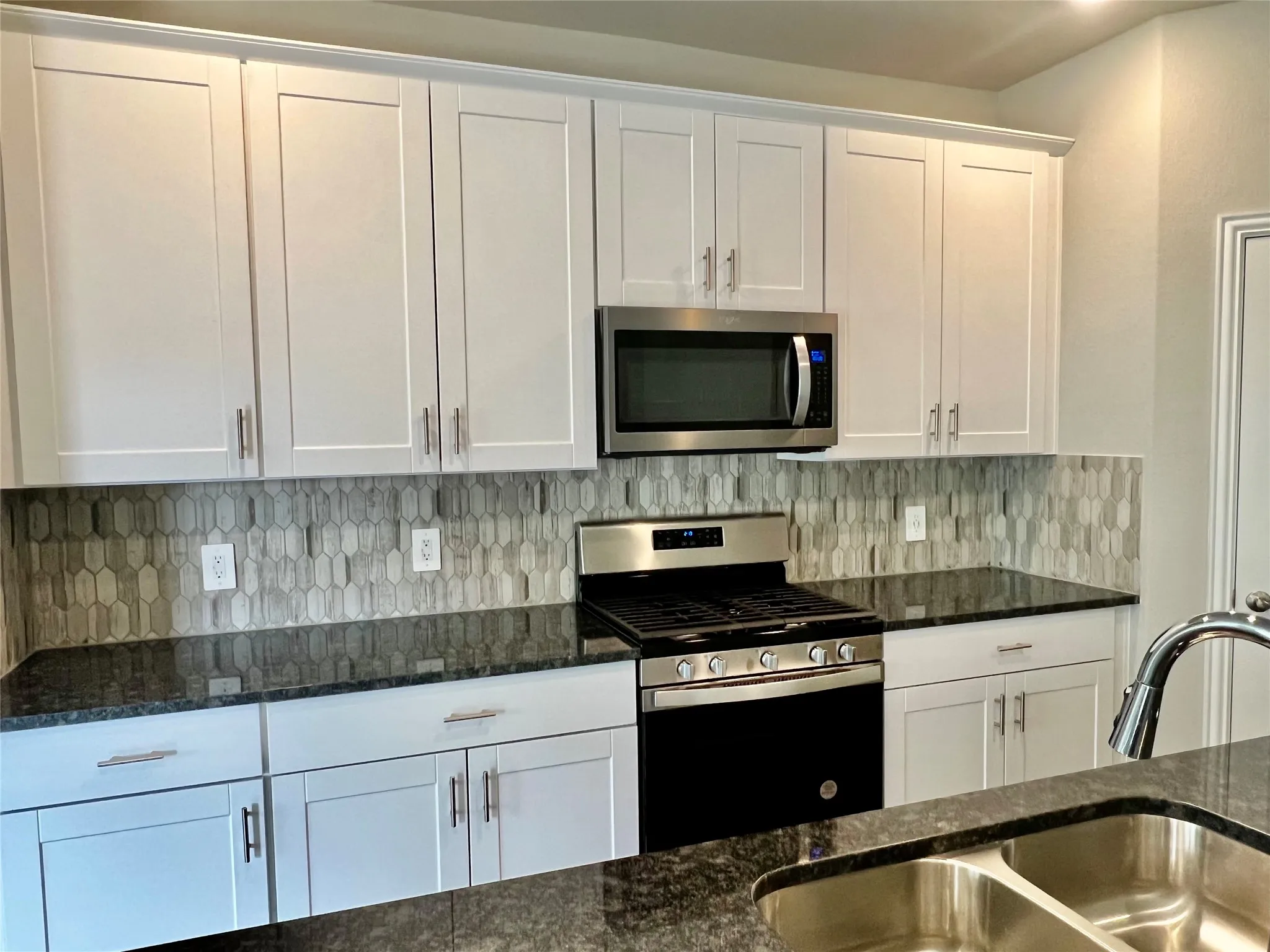 Kitchen featuring white shaker-style cabinetry, dark granite-finish countertops, an elongated hexagonal tile backsplash, stainless steel appliances, and a double basin sink with a chrome faucet