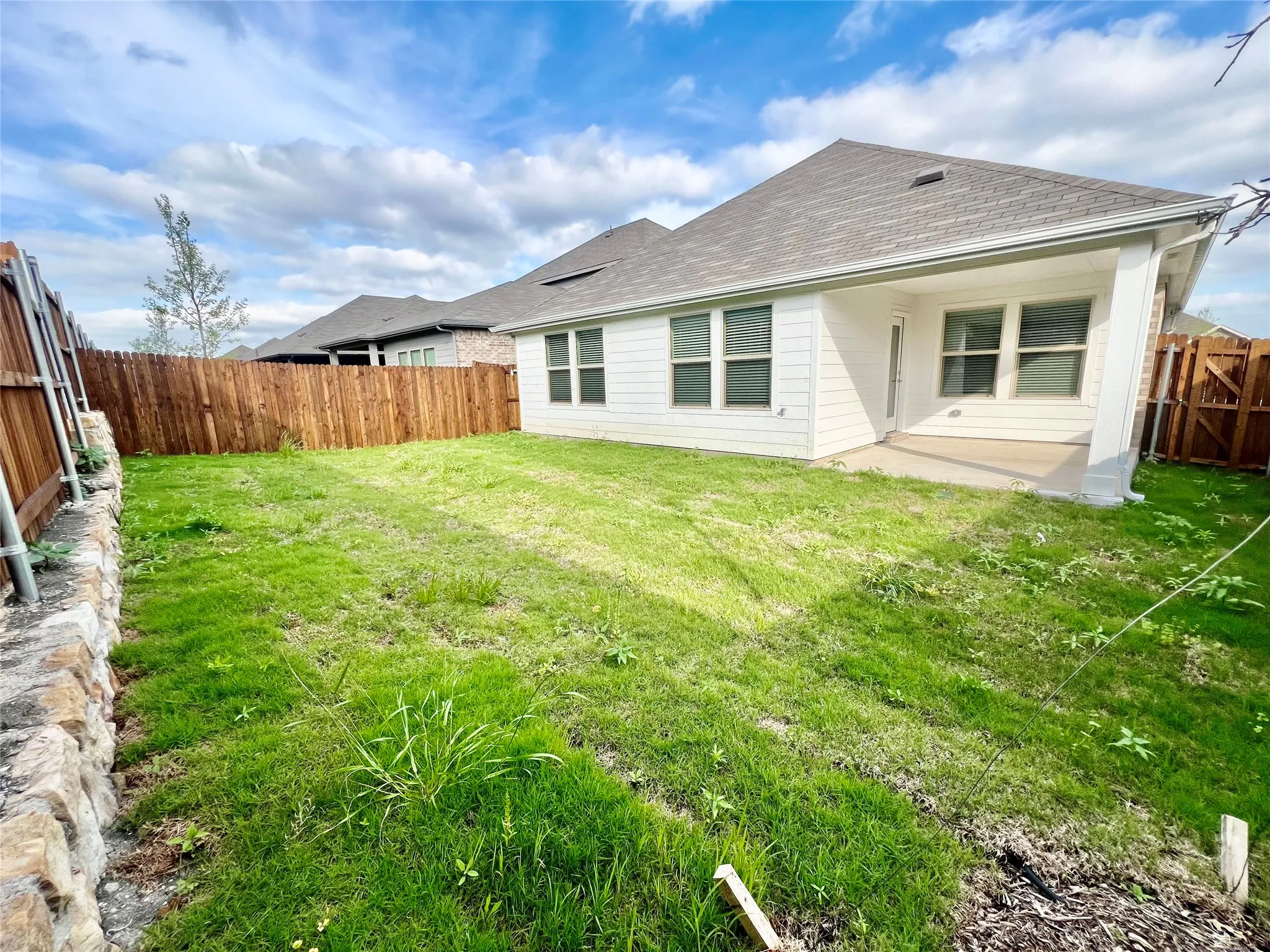 Expansive backyard featuring a covered patio, wood privacy fencing, a stone retaining wall, and a large lawn area