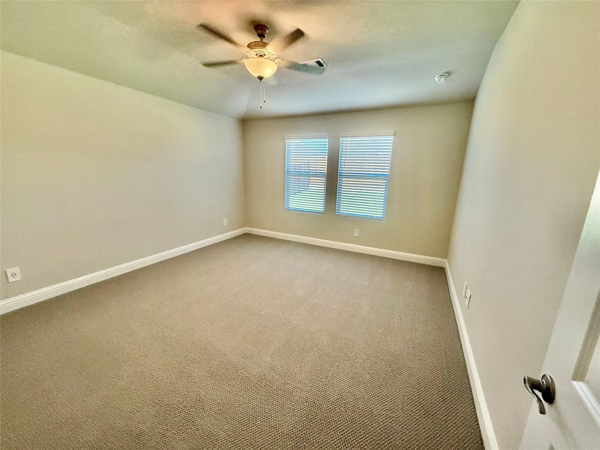 Spacious room featuring neutral carpeting, two windows with blinds, a ceiling fan with integrated lighting, and white baseboards