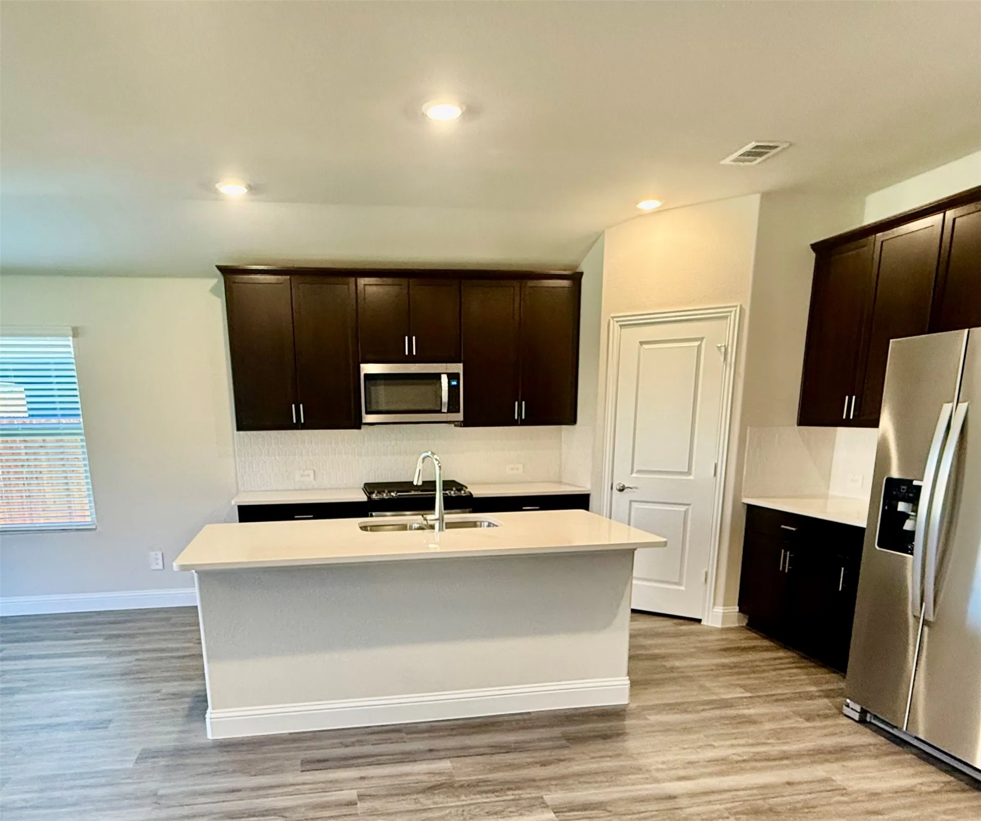 Modern kitchen featuring dark wood cabinetry, light countertops, stainless steel appliances, a center island with sink, and wood-finish flooring