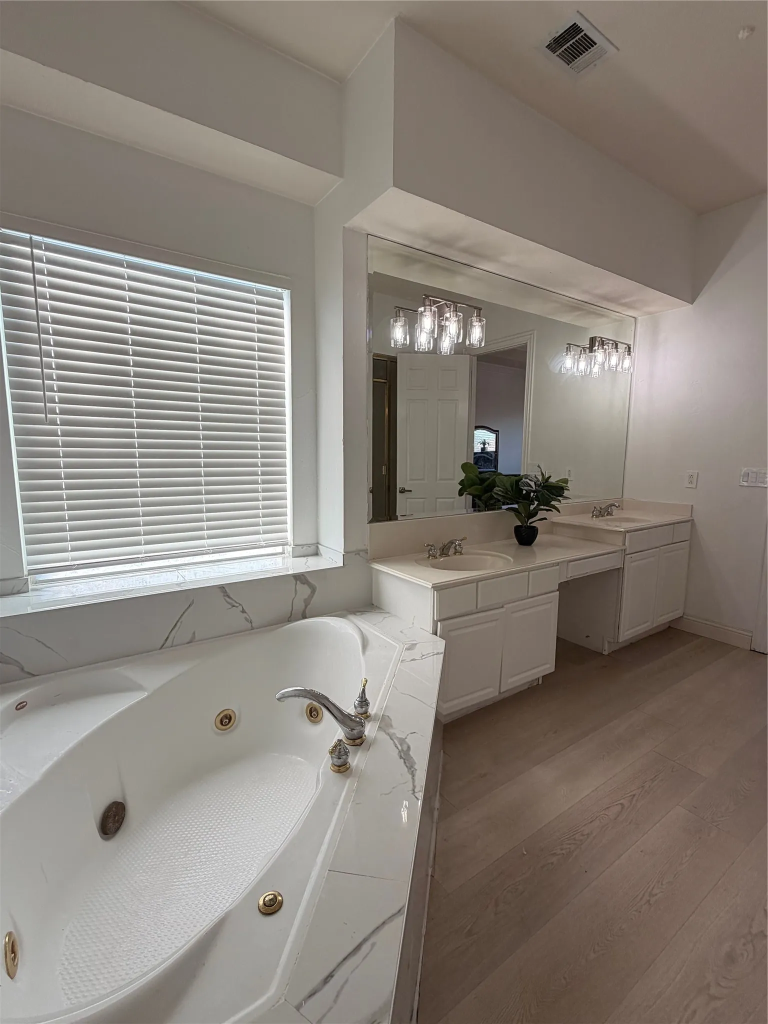 Bathroom featuring a built-in jetted tub with a marble-look surround, dual vanity with undermount sinks, and wood-finish flooring