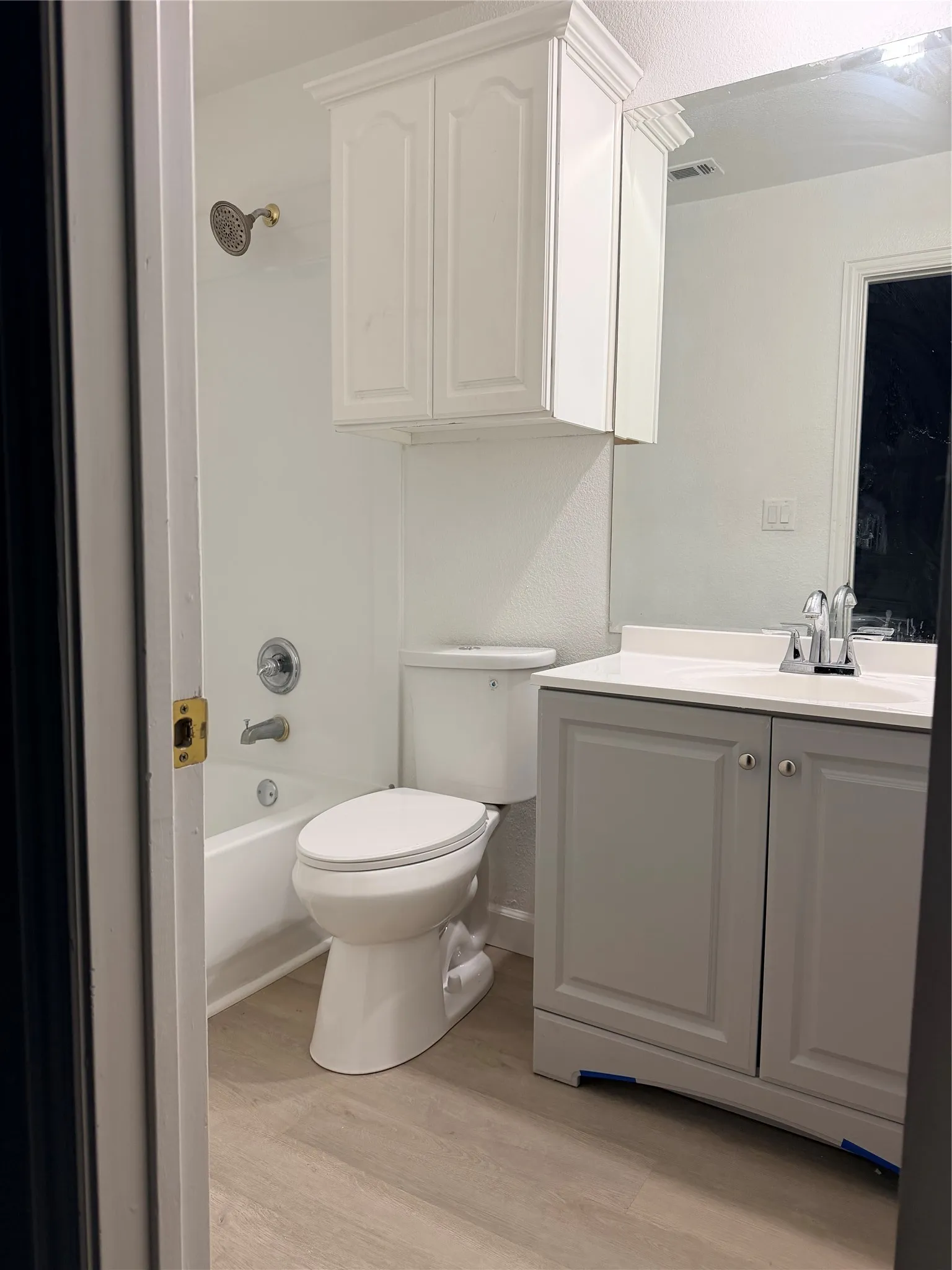 Bathroom featuring a white tub and shower combination with a decorative showerhead, a white toilet, a white upper cabinet with crown molding, a vanity with gray shaker-style doors, and wood-finish flooring