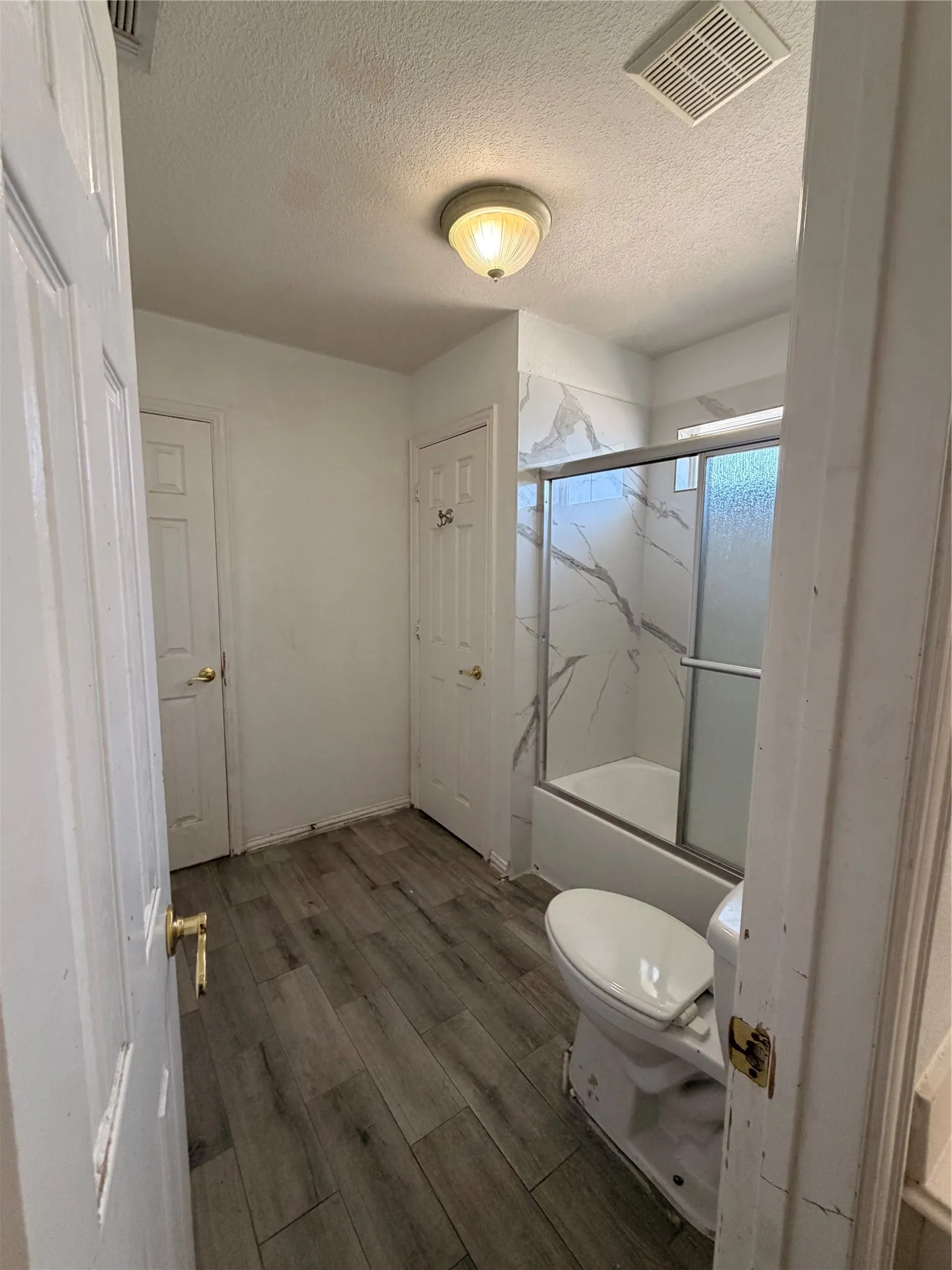 Bathroom featuring a shower/tub combination with patterned tile surround and sliding glass doors, a white toilet, and wood-finish flooring