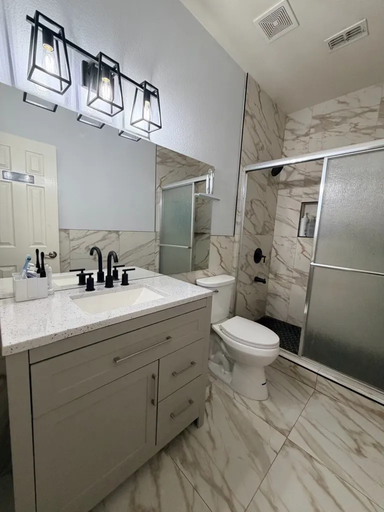 Bathroom featuring a white vanity with a light speckled countertop, matte black fixtures, a shower with sliding glass doors and black hexagon tile flooring, and large format marbled wall and floor tiles