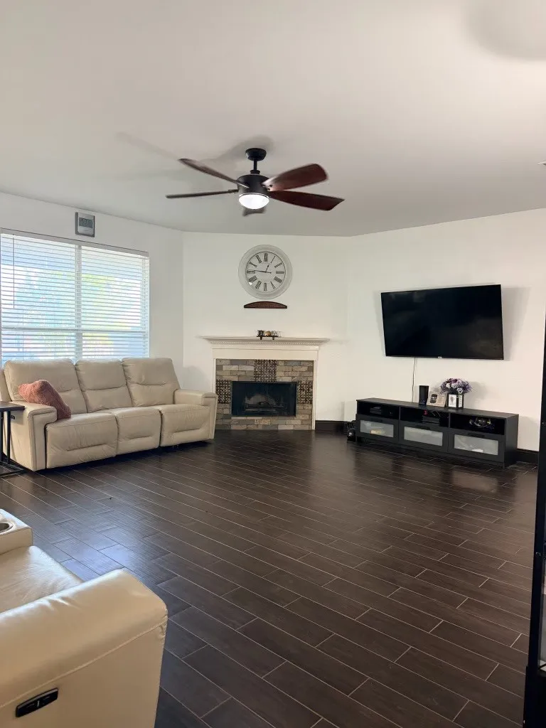 Living room featuring wood-finish flooring, a corner fireplace with stone surround and mantle, and a ceiling fan with integrated lighting