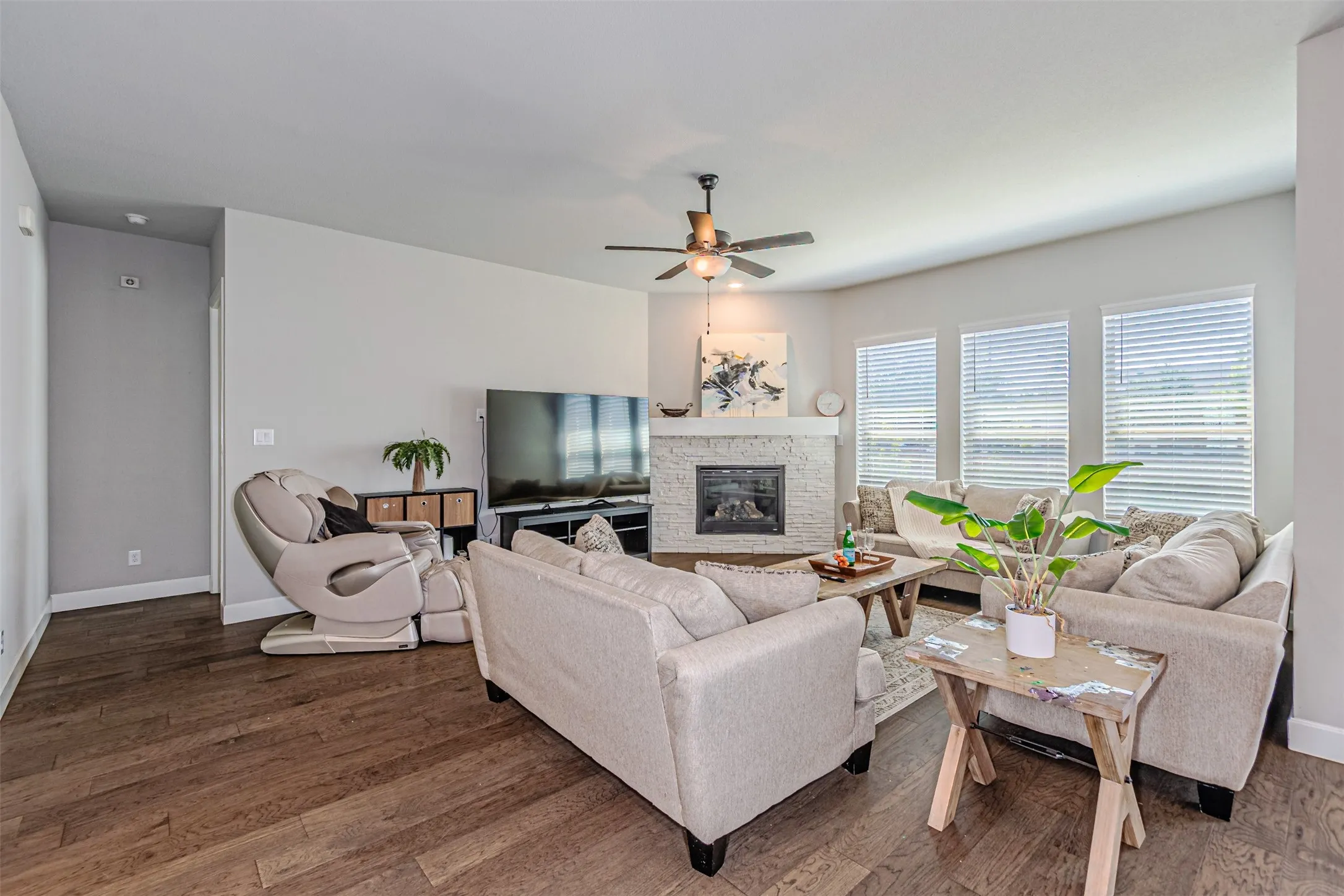 Expansive living area with wood-finish flooring, a corner fireplace featuring stone surround, multiple windows with blinds, and an overhead ceiling fan with lighting