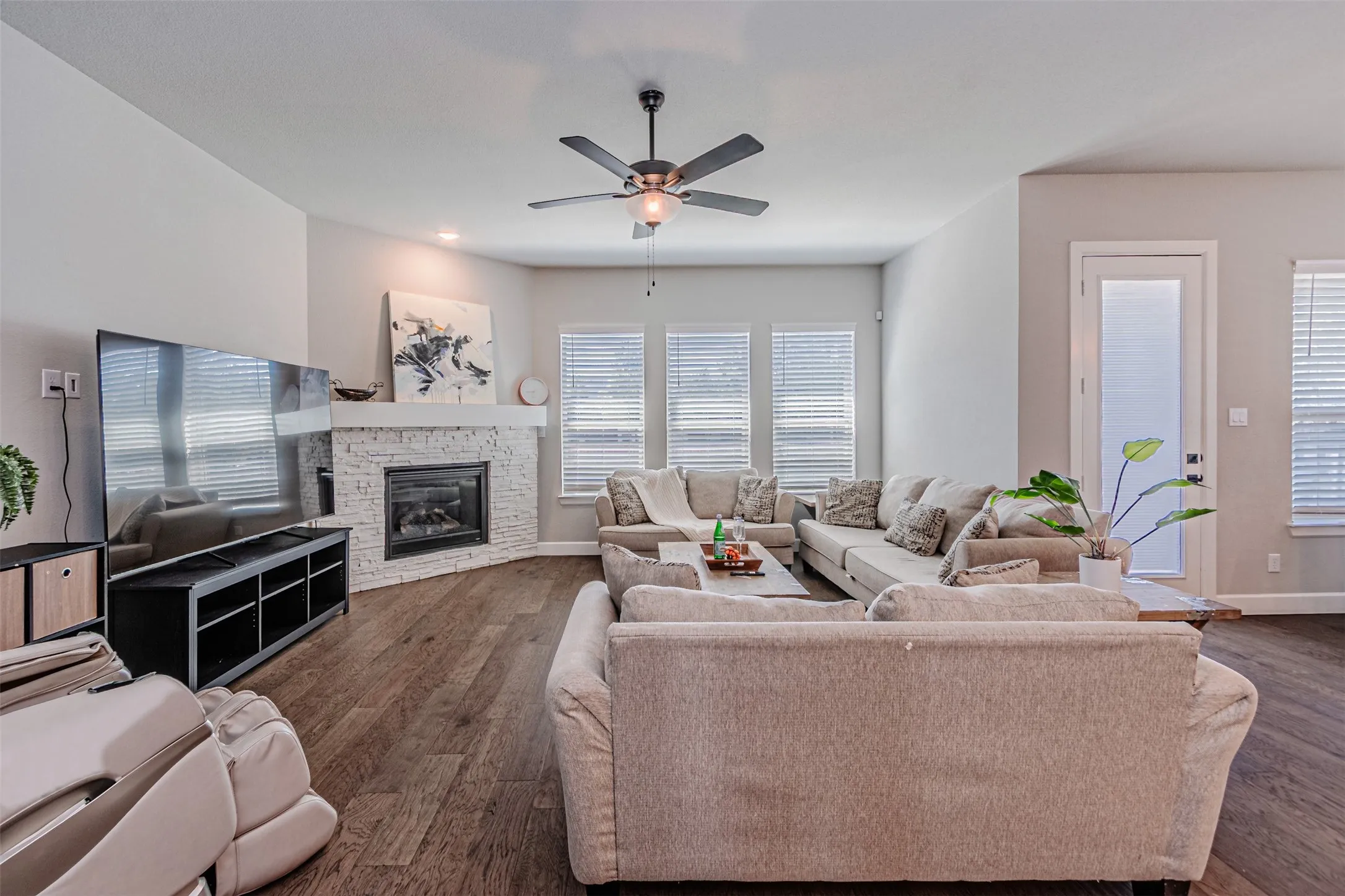 Wood-finish flooring, corner fireplace with white stacked stone surround, ceiling fan, and multiple windows with blinds