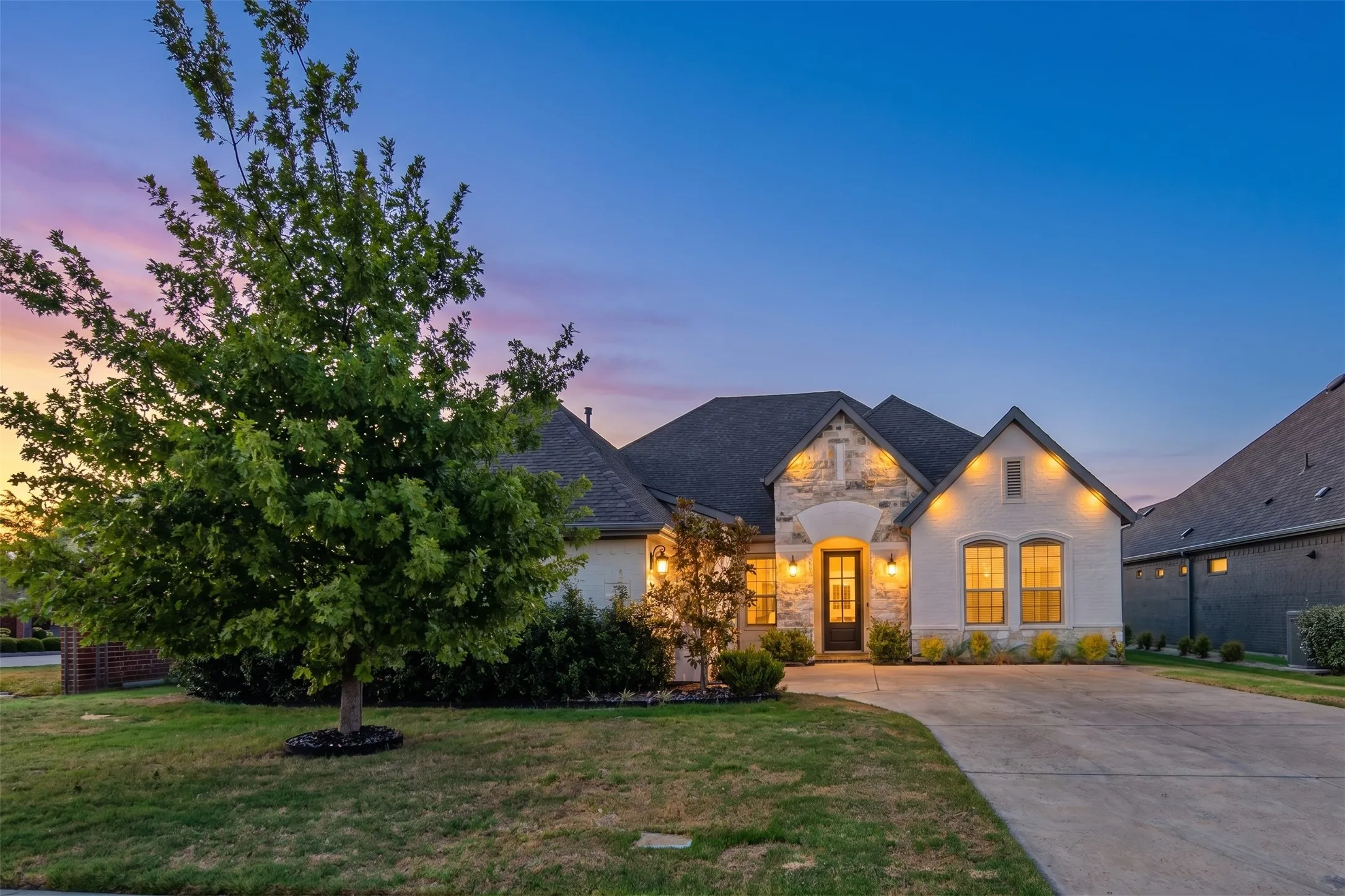 Stone and stucco facade, gabled roofline, concrete driveway, and established landscaping