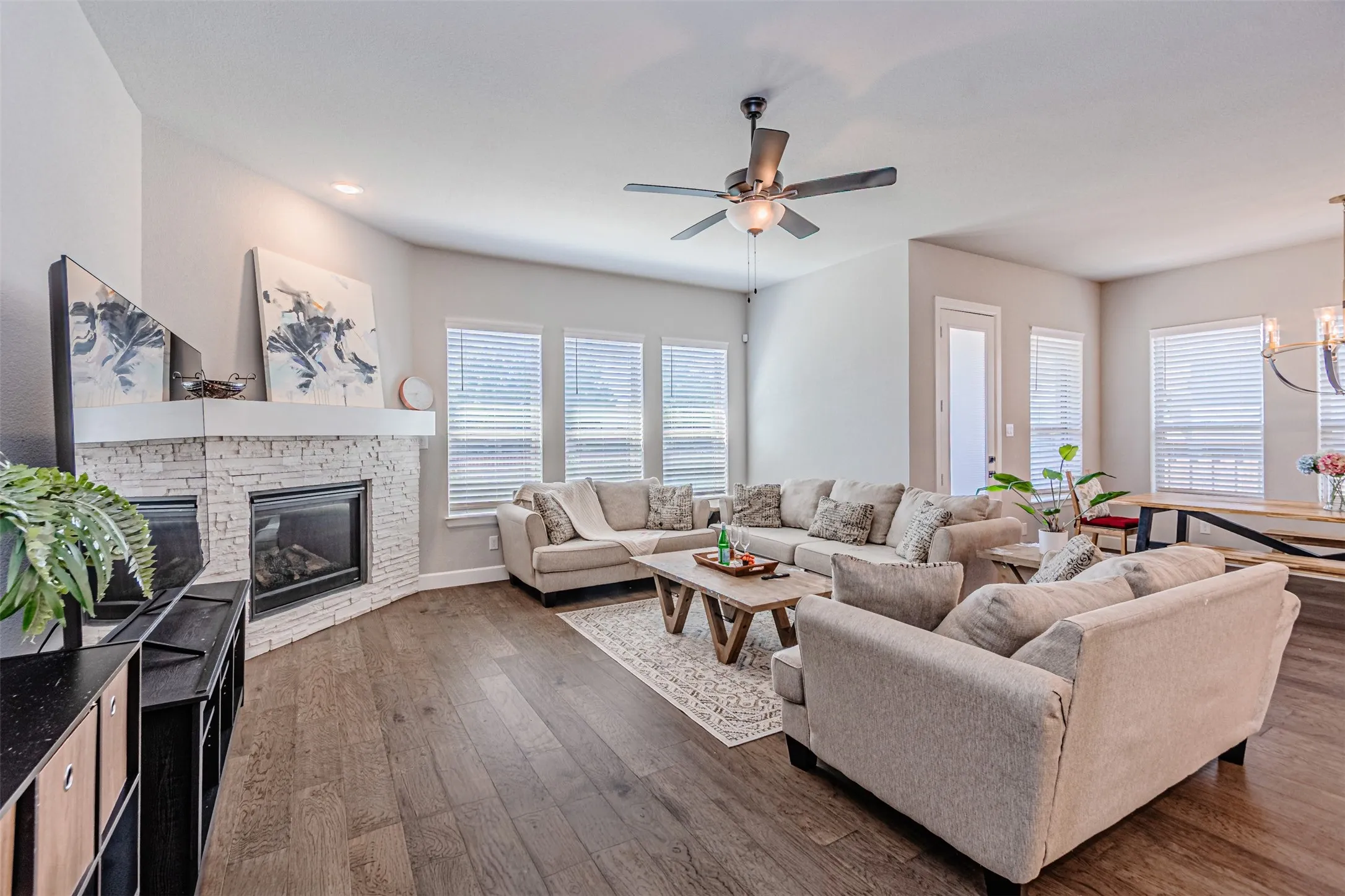 Open-concept living area featuring wood-finish flooring, a corner fireplace with a white stone surround, multiple windows with blinds, and a ceiling fan