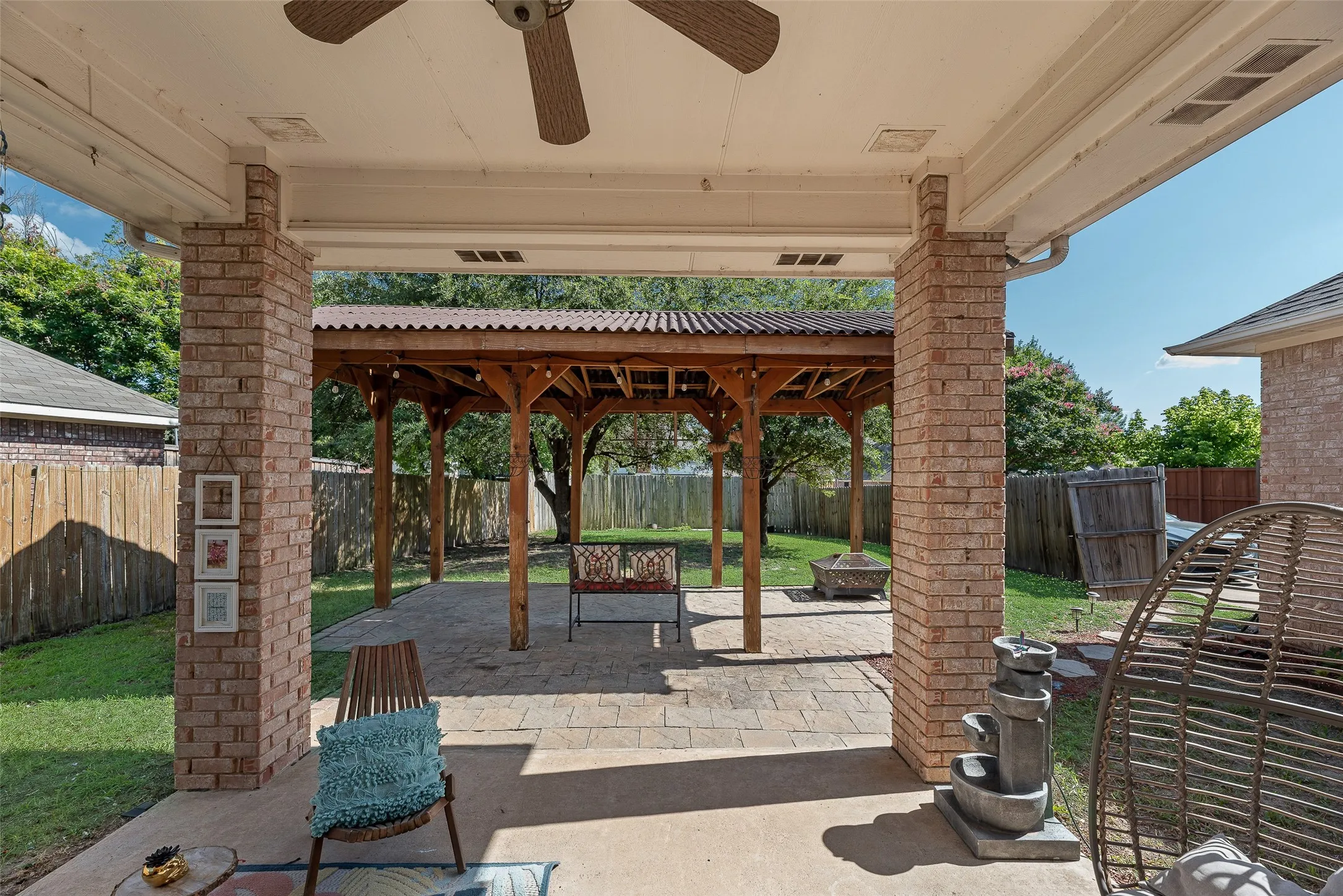 Covered patio with brick columns and ceiling fan, extending to a paver patio with a wood-framed pergola and corrugated metal roof