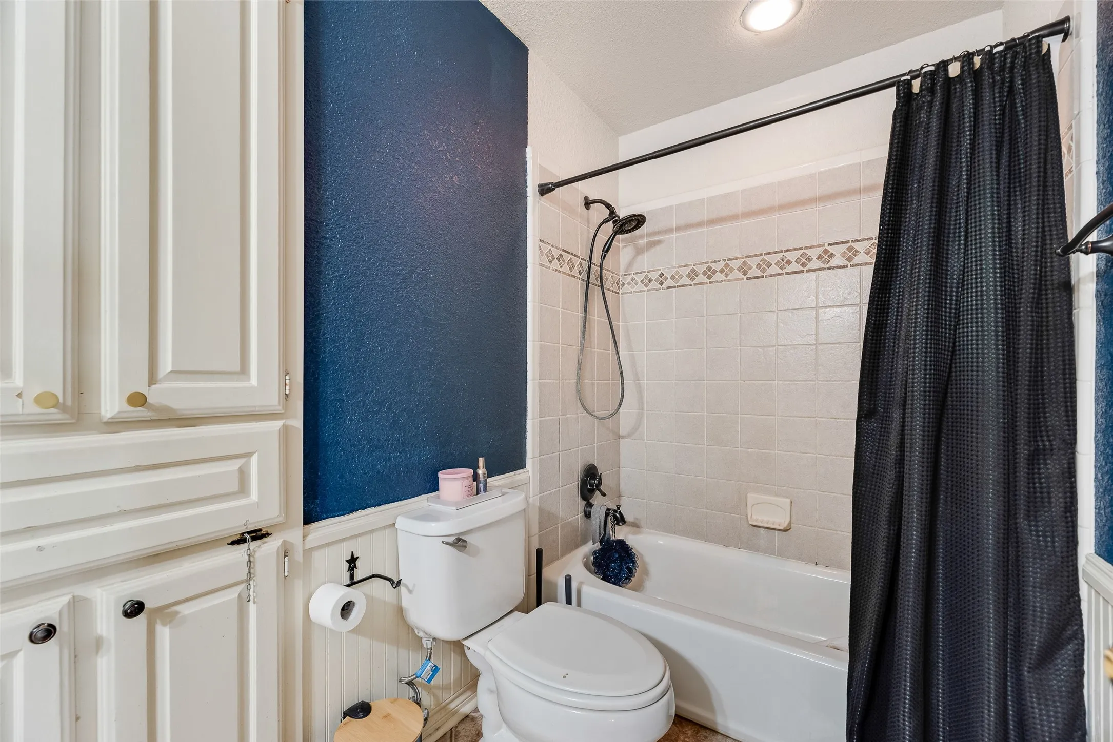 Bathroom featuring a white tub, shower with ceramic tile surround and decorative accent tile, built-in white cabinetry, and a toilet with beadboard wainscoting
