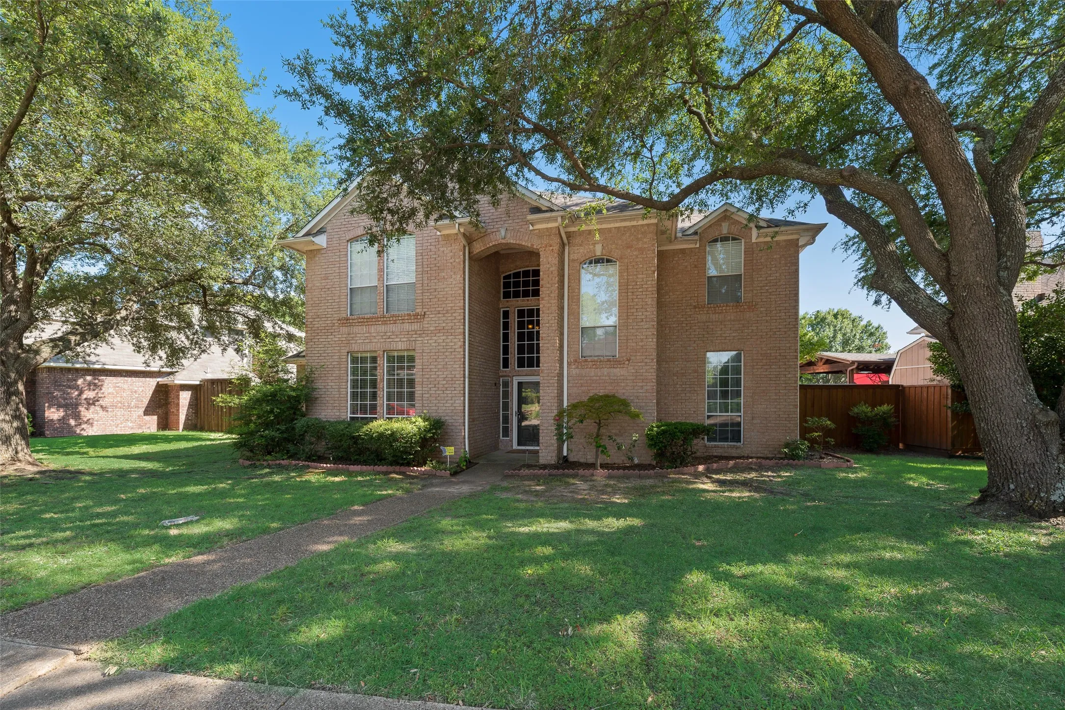 Two-story brick facade residence featuring a prominent arched entry, multiple large windows, a well-maintained lawn, mature trees, and established shrubbery