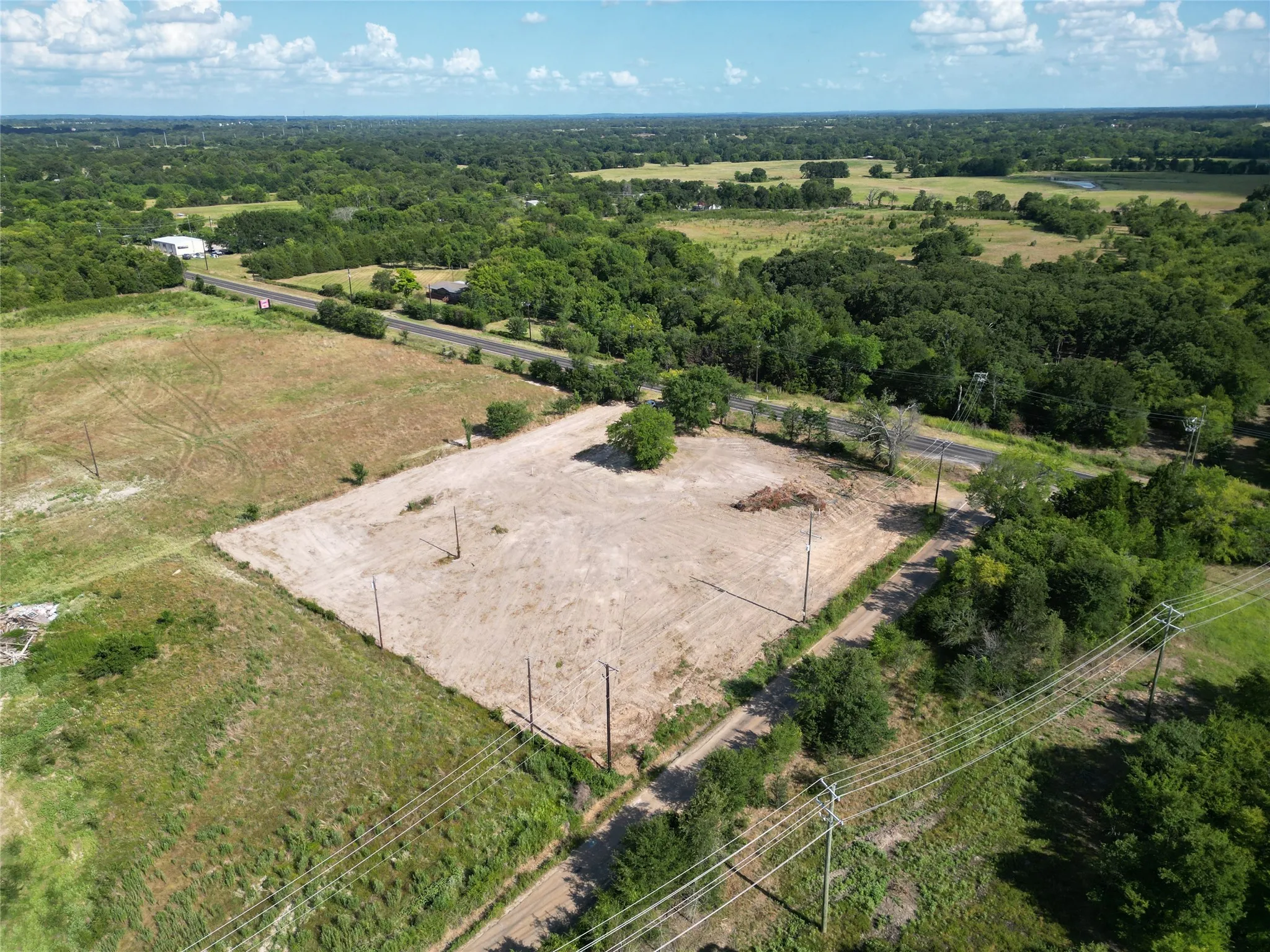 Expansive cleared lot bordered by mature trees and a paved road, featuring power lines and a distant building