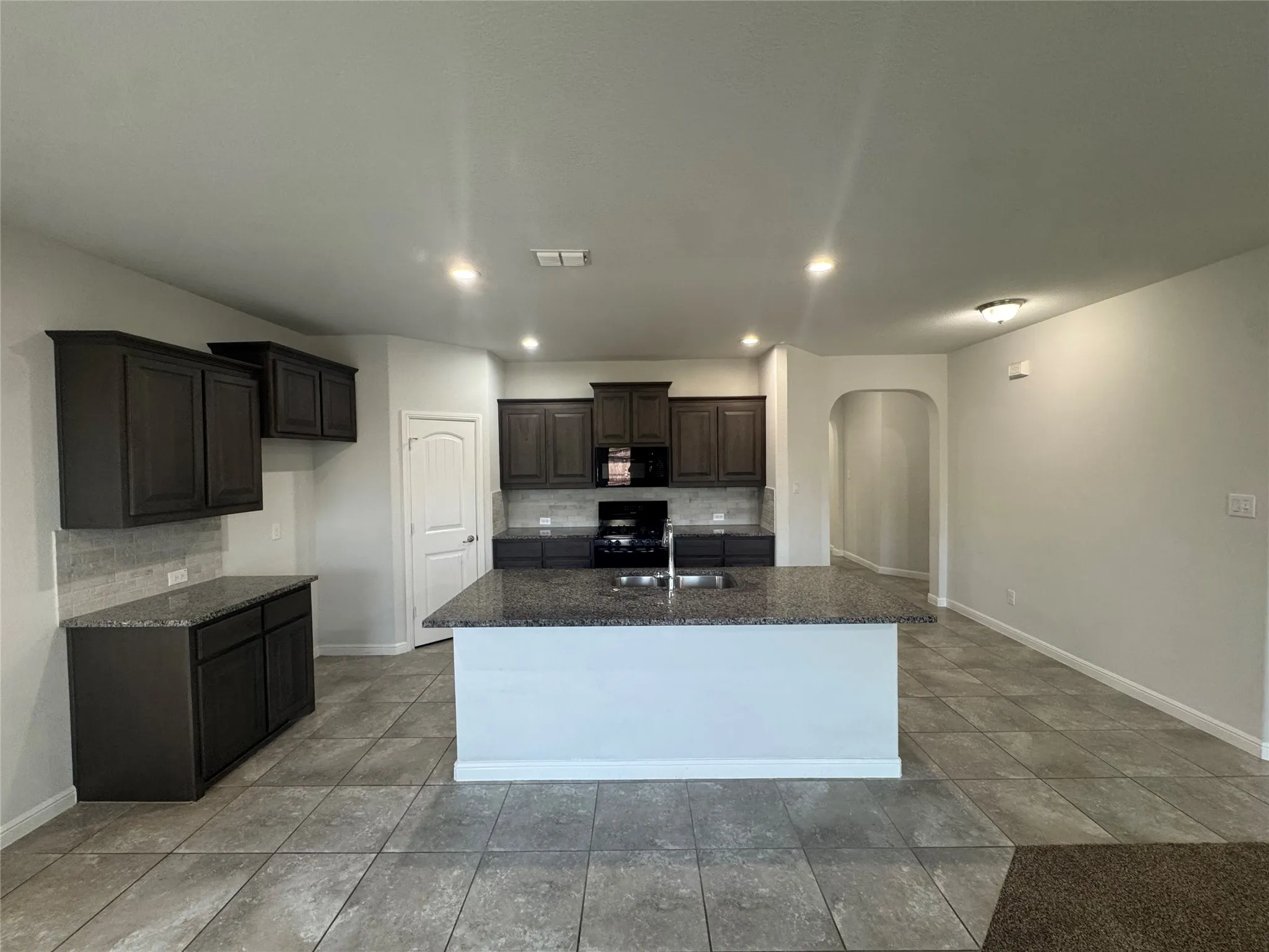 Kitchen featuring dark wood cabinetry, granite countertops, tile backsplash, built-in appliances, and a central island with an integrated sink