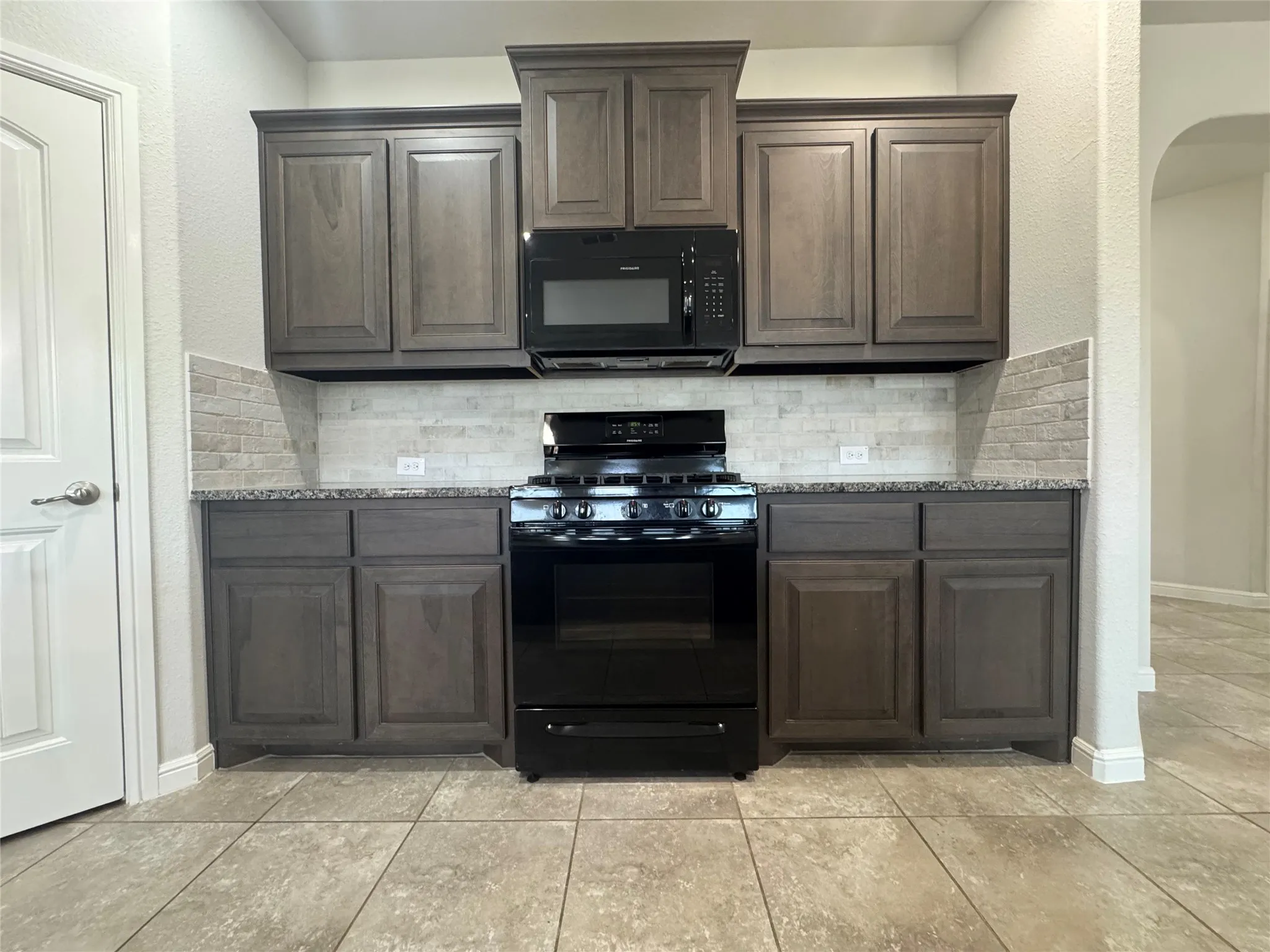 Stove with overhead microwave, natural stone backsplash, dark wood-finish cabinetry, and granite-style countertops