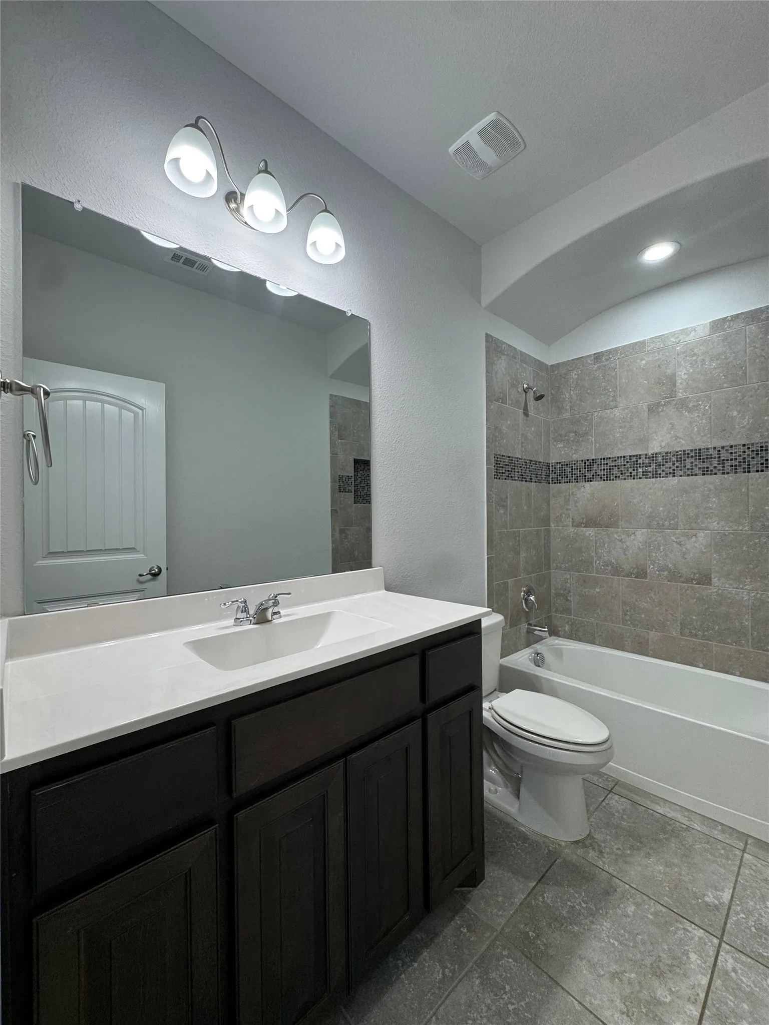 White porcelain bathtub with tile surround, dark wood-finish vanity with white countertop, and tiled flooring