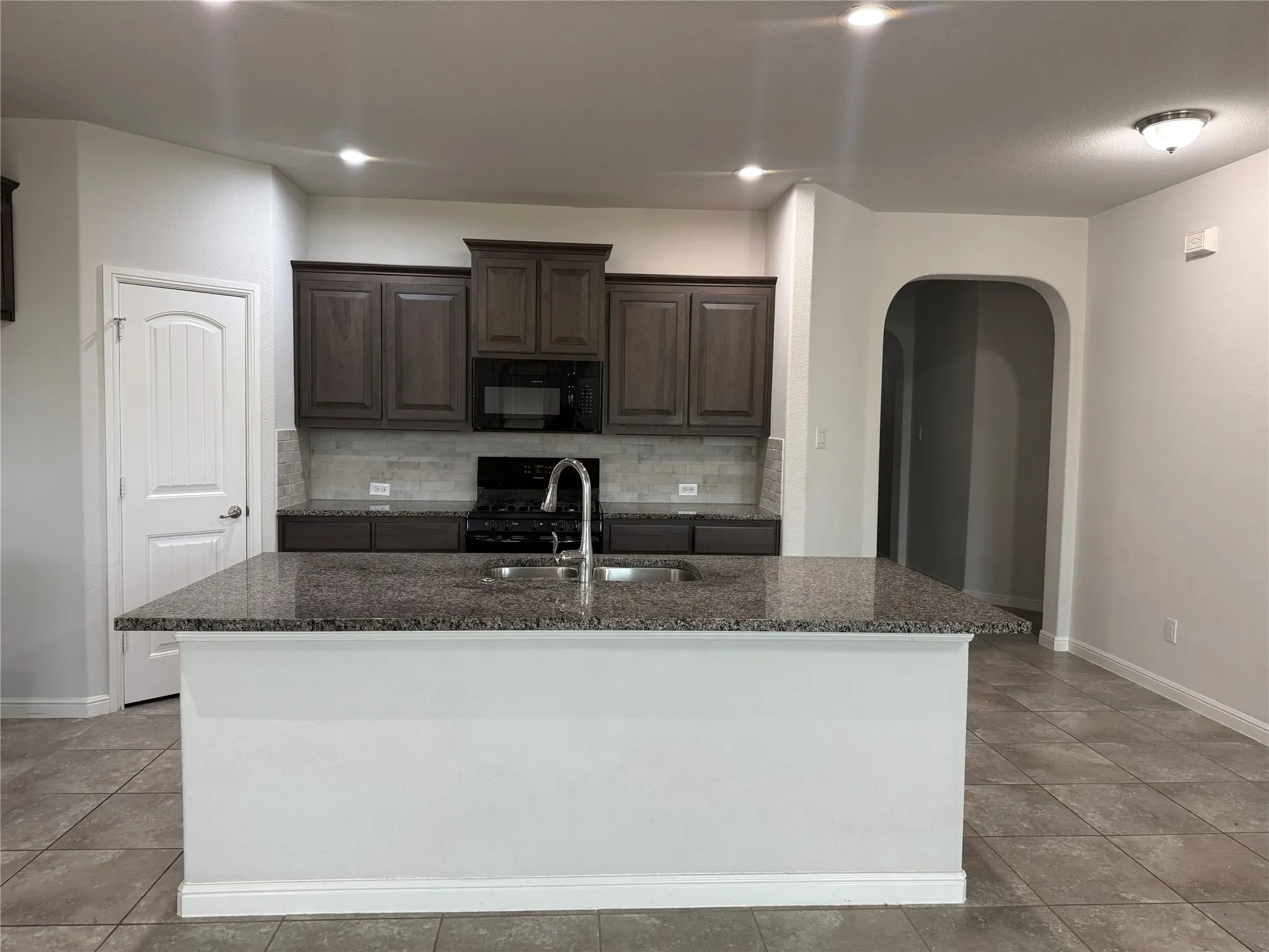 Kitchen featuring a central island with a sink and granite countertop, dark wood-finish cabinetry, a black appliance suite, and a tiled backsplash