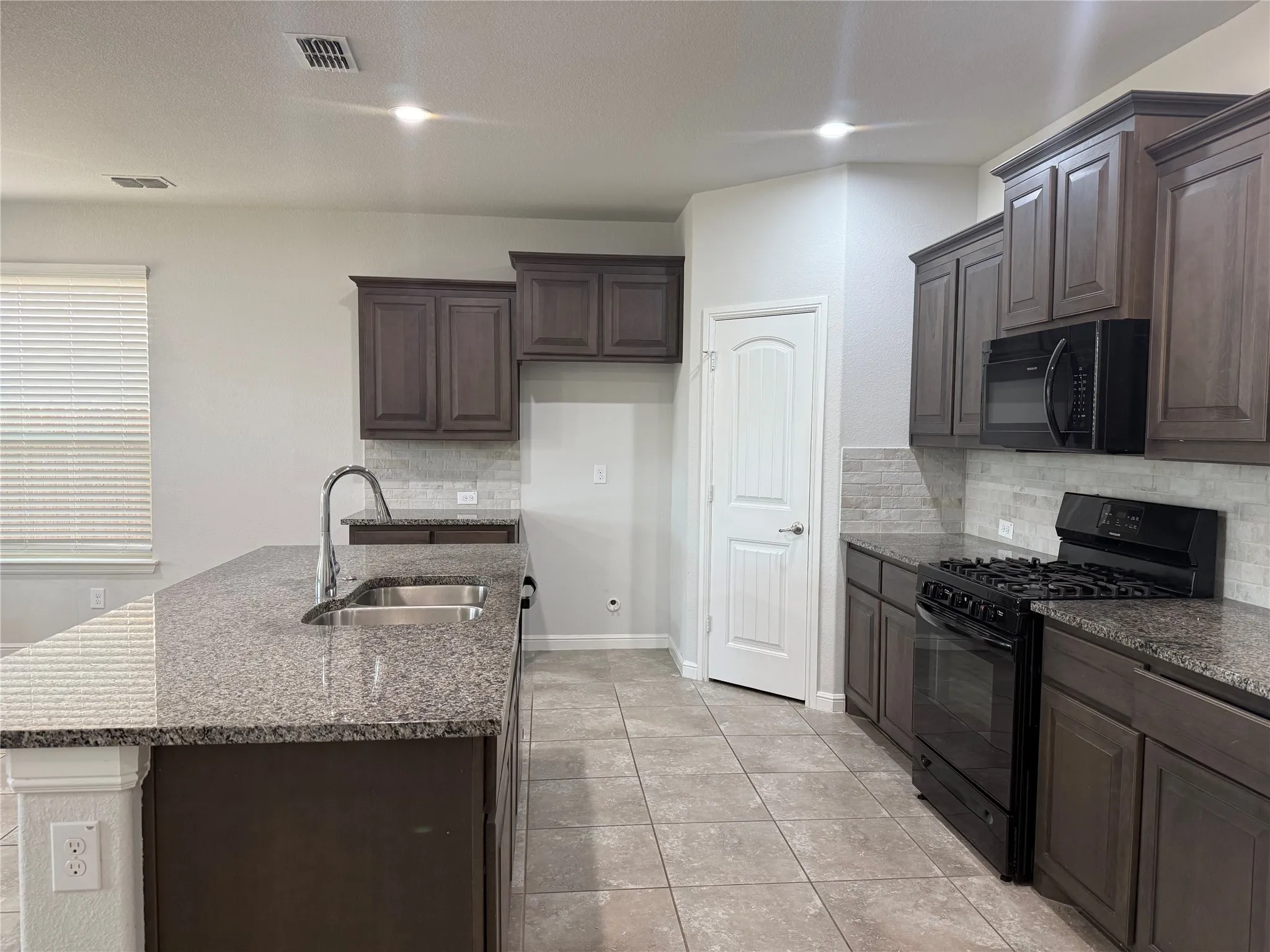 Kitchen featuring a central island with an undermount double-basin sink, granite countertops, dark wood cabinetry, a tiled backsplash, and tile flooring