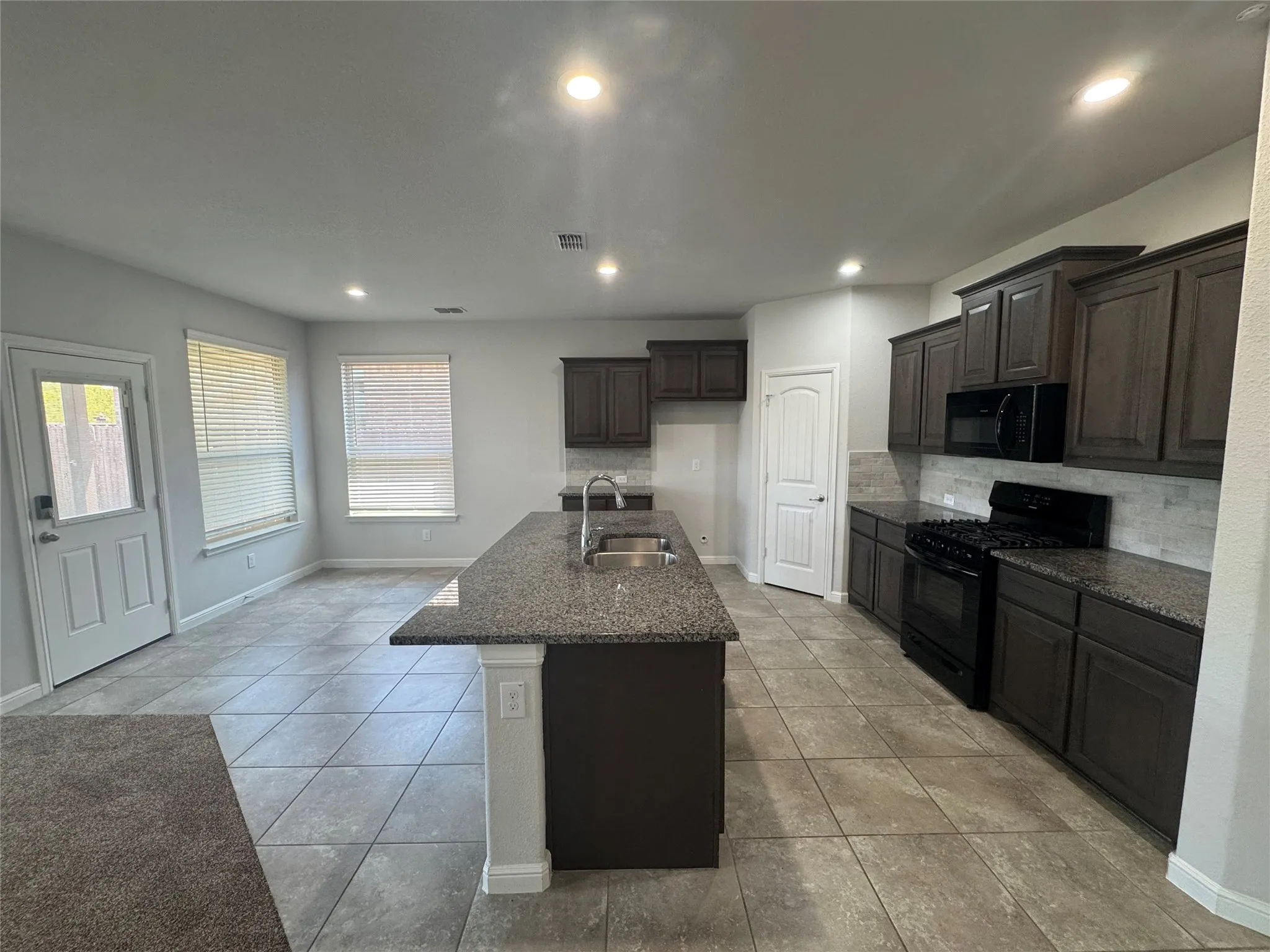 Open-concept kitchen featuring dark wood-finish cabinetry, granite countertops, a central island with an undermount sink, a subway tile backsplash, and tile flooring