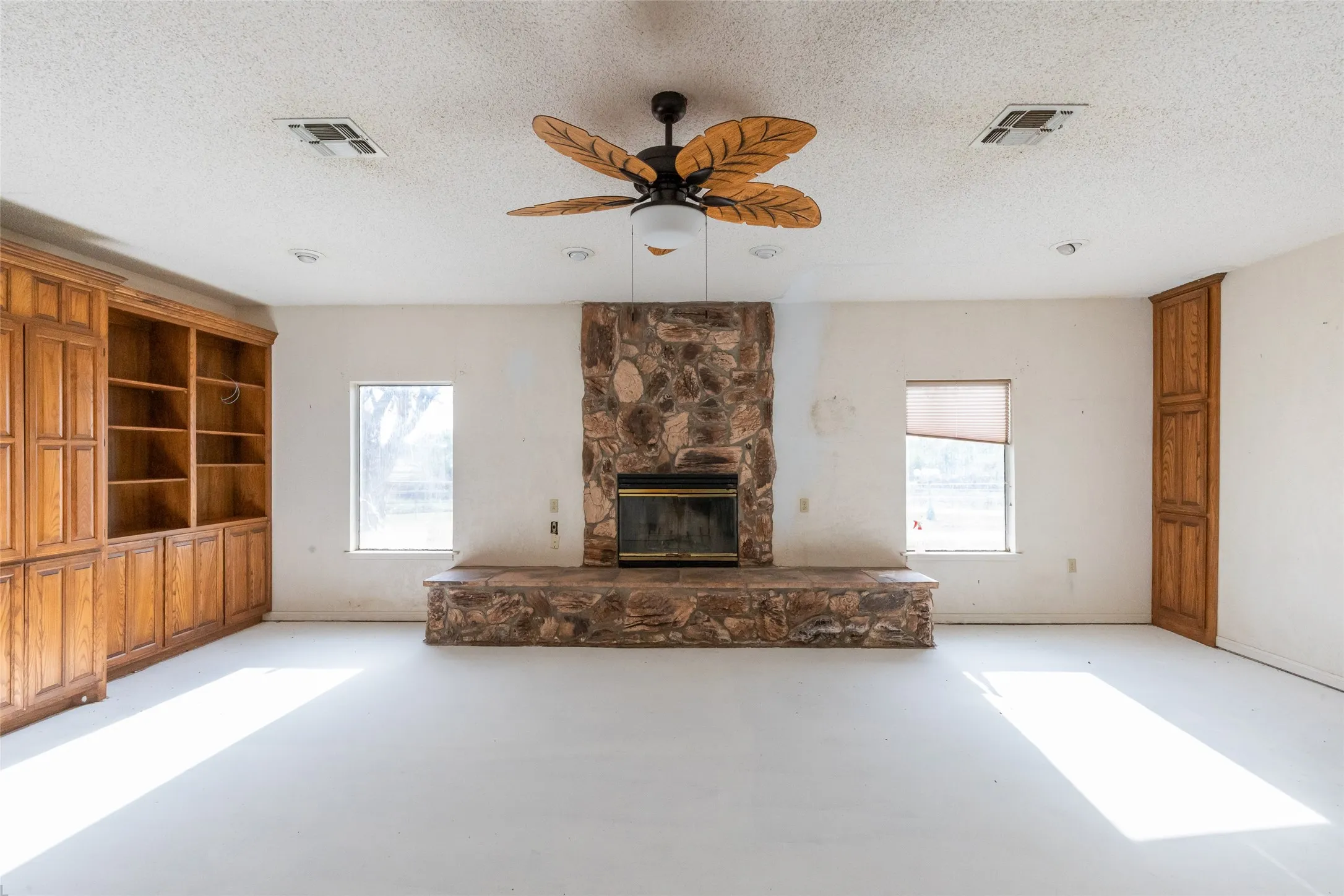 Spacious room featuring a stone-clad fireplace with a stone hearth, built-in wooden cabinetry with open shelving, additional built-in wooden storage, recessed lighting, and a ceiling fan with leaf-shaped blades