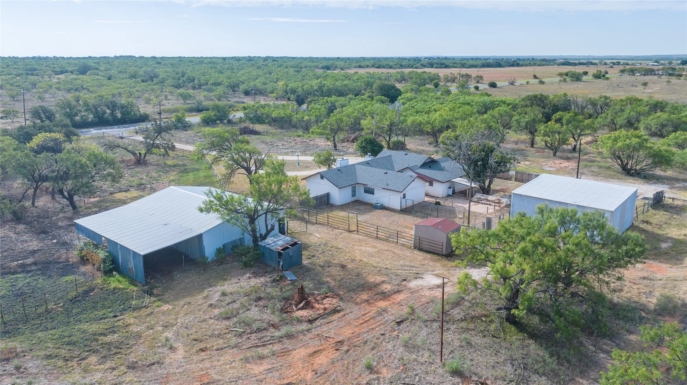 Expansive rural property featuring a white-sided residence with dark roofing, multiple outbuildings including a large metal barn and a spacious metal workshop, and mature trees throughout the acreage