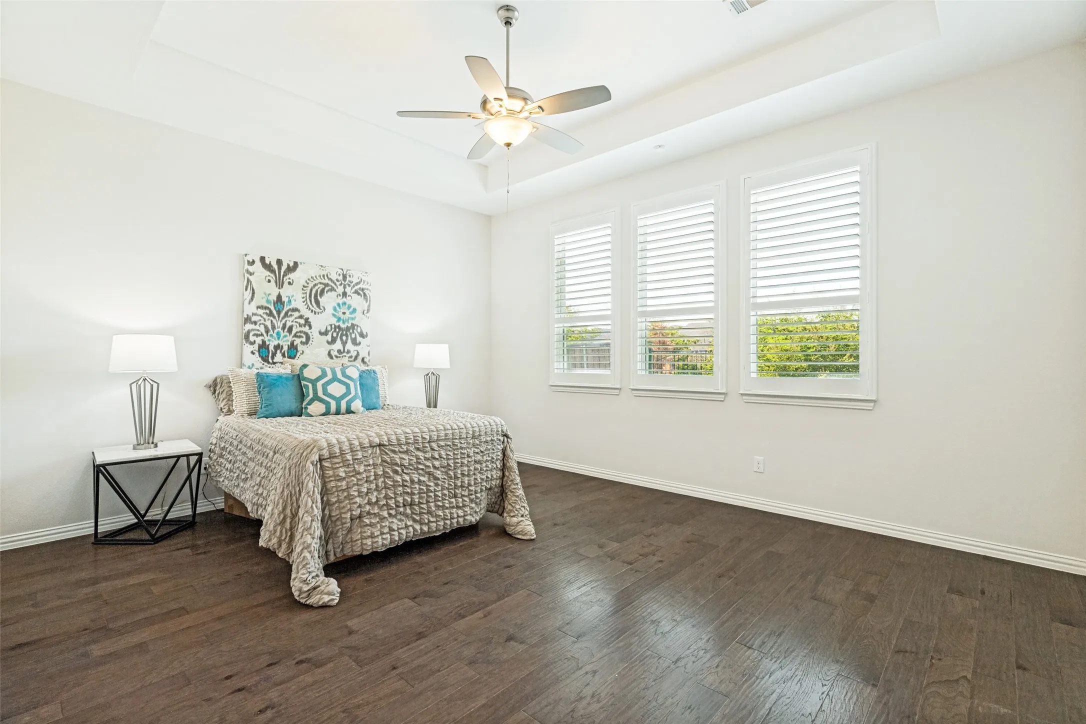 Spacious room featuring dark wood-finish flooring, three windows with plantation shutters, a tray ceiling, and a ceiling fan with integrated lighting