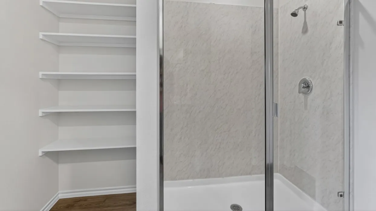 Built-in shelving unit with white shelves, adjacent to a shower enclosure featuring a clear glass door, chrome fixtures, and textured shower surround panels
