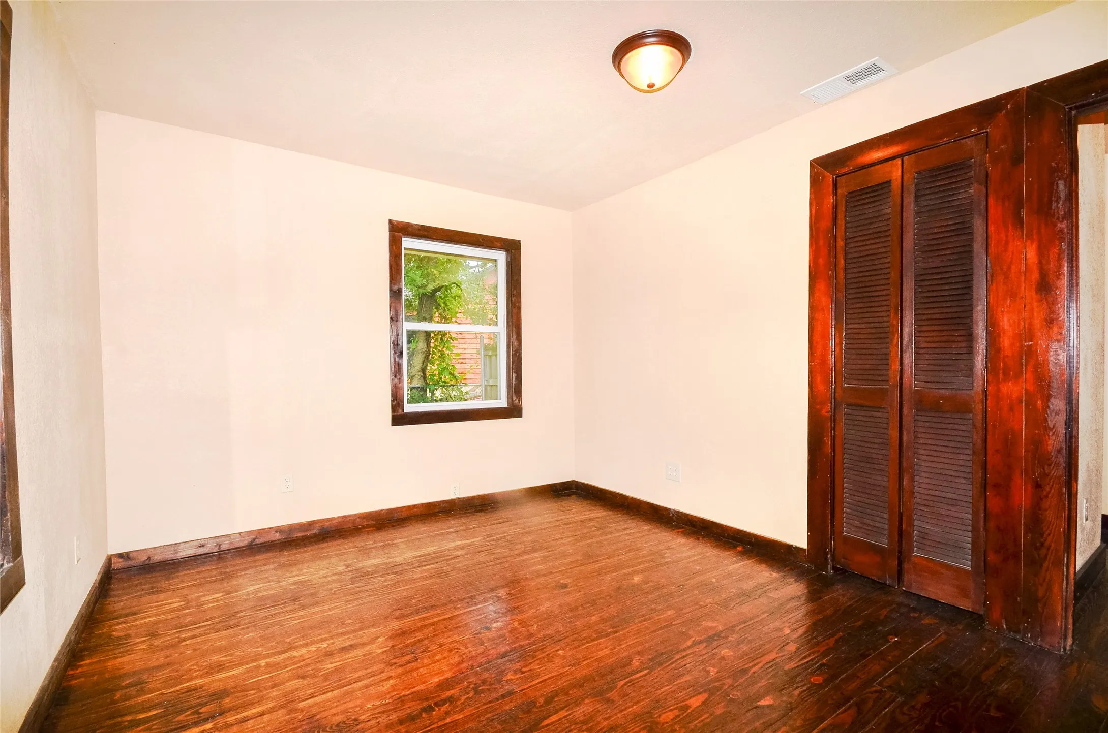 Room featuring wood-finish flooring, a single window with wood trim, louvered bi-fold doors, and a flush-mount ceiling light fixture