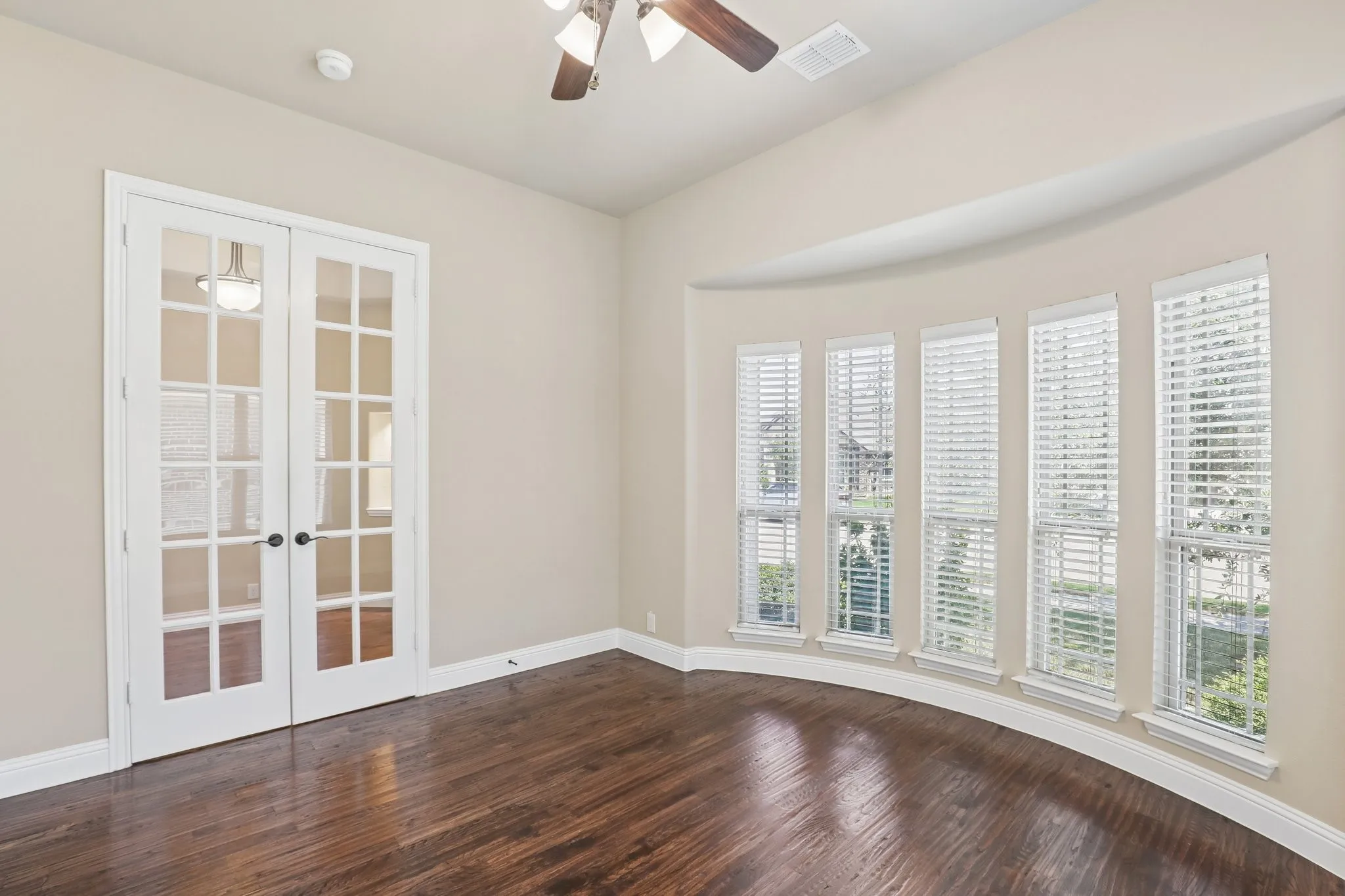 Room featuring dark wood-finish flooring, a curved wall with five vertical windows, white window blinds, and white French doors