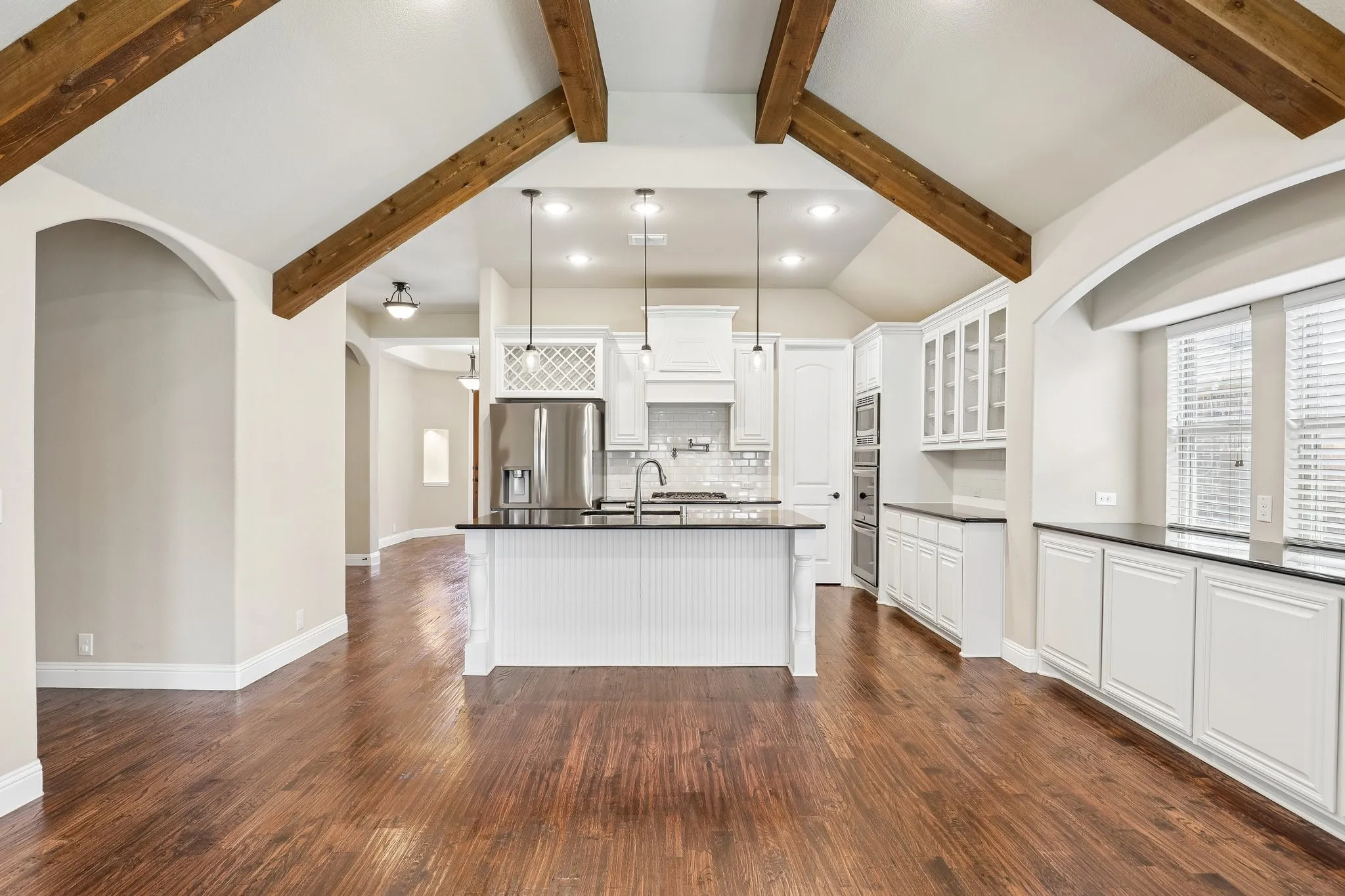 Open-concept kitchen and living area featuring exposed wood ceiling beams, rich wood-finish flooring, a central island with dark countertops, white cabinetry, and stainless steel appliances