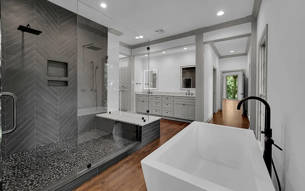 Modern bathroom featuring a freestanding soaking tub with a matte black floor-mounted faucet, a spacious glass-enclosed shower with dark chevron-patterned tile, and a dual vanity with light gray cabinetry and integrated sinks