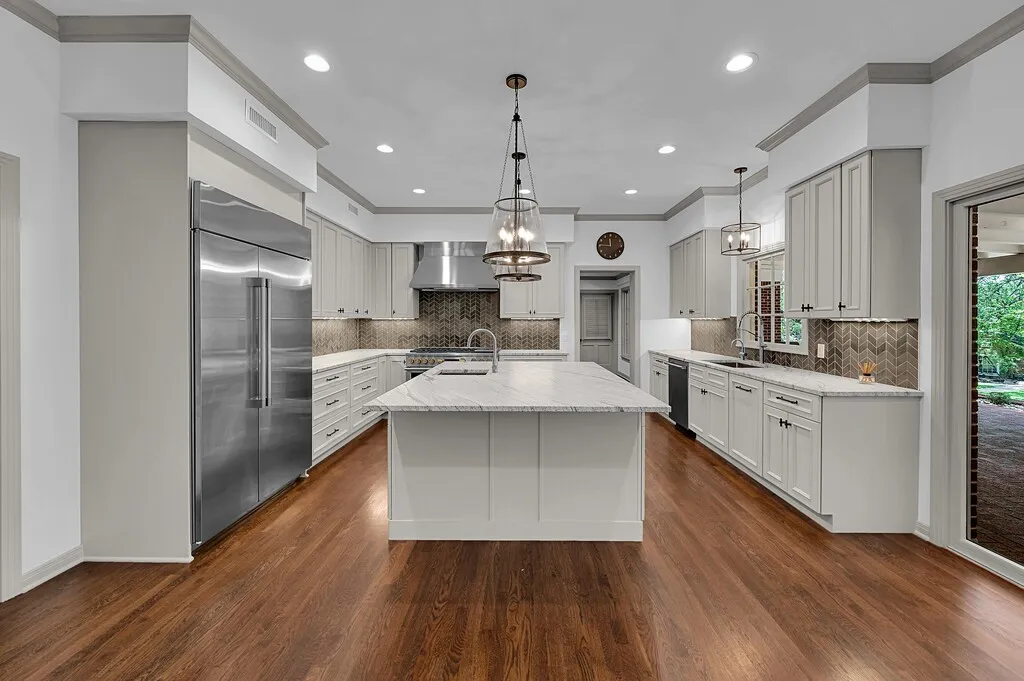Spacious kitchen featuring a central island with a light-toned countertop, stainless steel appliances, light-toned cabinetry, a herringbone pattern backsplash, and wood-finish flooring
