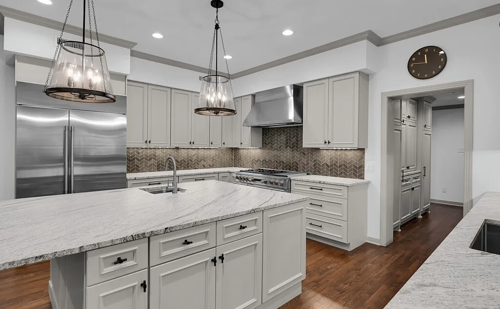 Gourmet kitchen featuring a large central island with an undermount sink, stainless steel appliances, light-toned cabinetry with dark hardware, a herringbone pattern backsplash, and wood-finish flooring