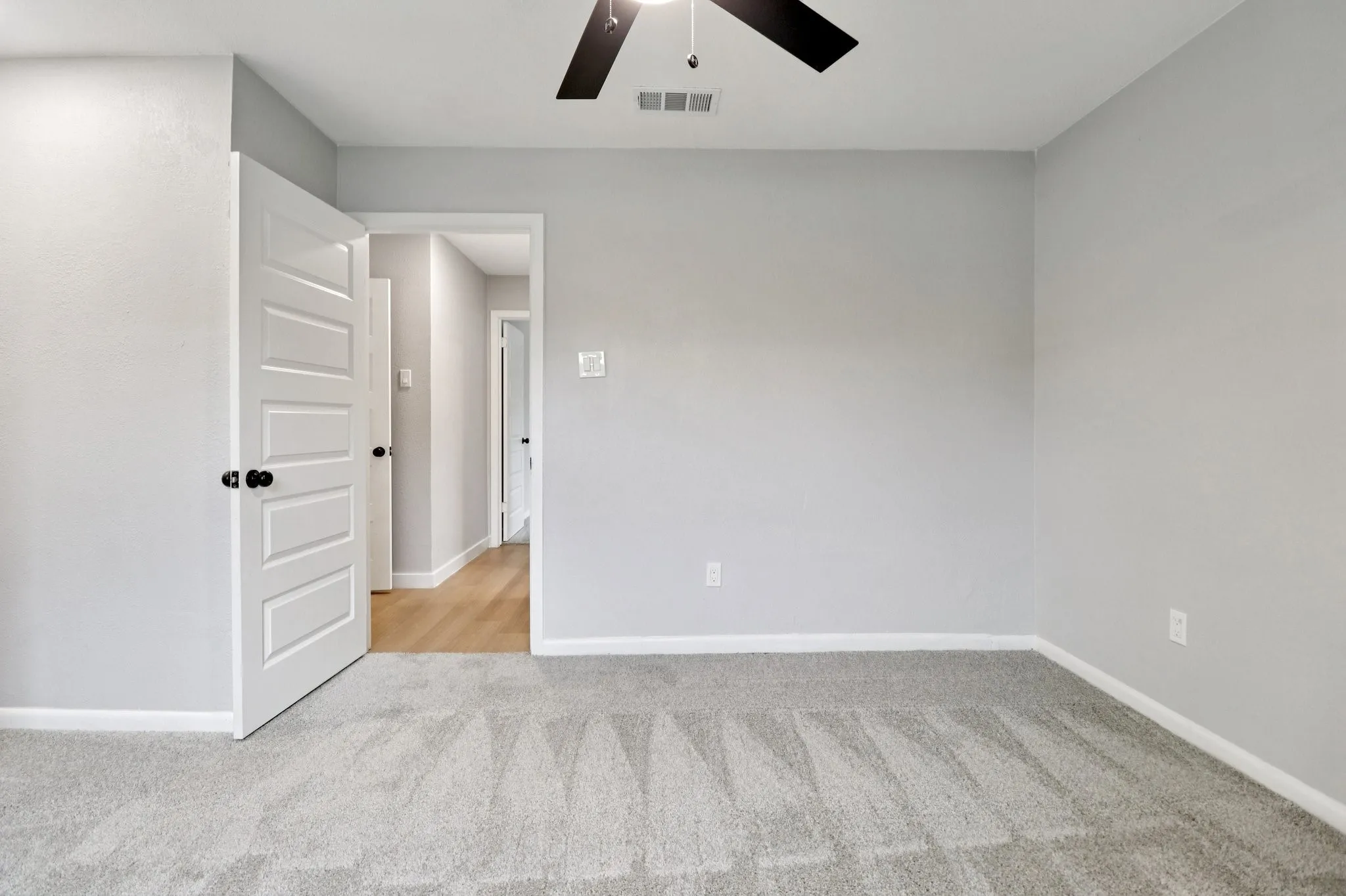 Interior room featuring light gray wall paint, gray carpeting, a white five-panel door with black hardware, and a ceiling fan with black blades